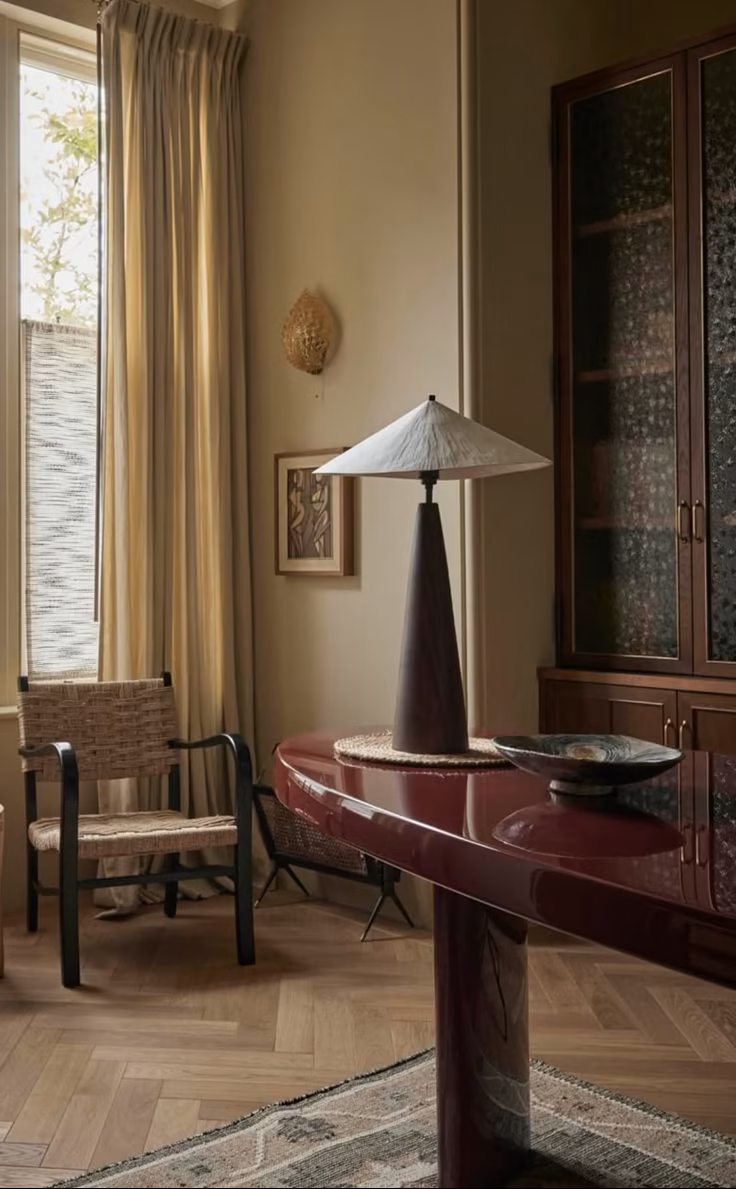 An elegant study with a burgundy lacquered desk, a modernist table lamp, a vintage woven wicker chair, a window with tall draped curtains, a framed picture on the wall, and a wooden cabinet with glass doors.