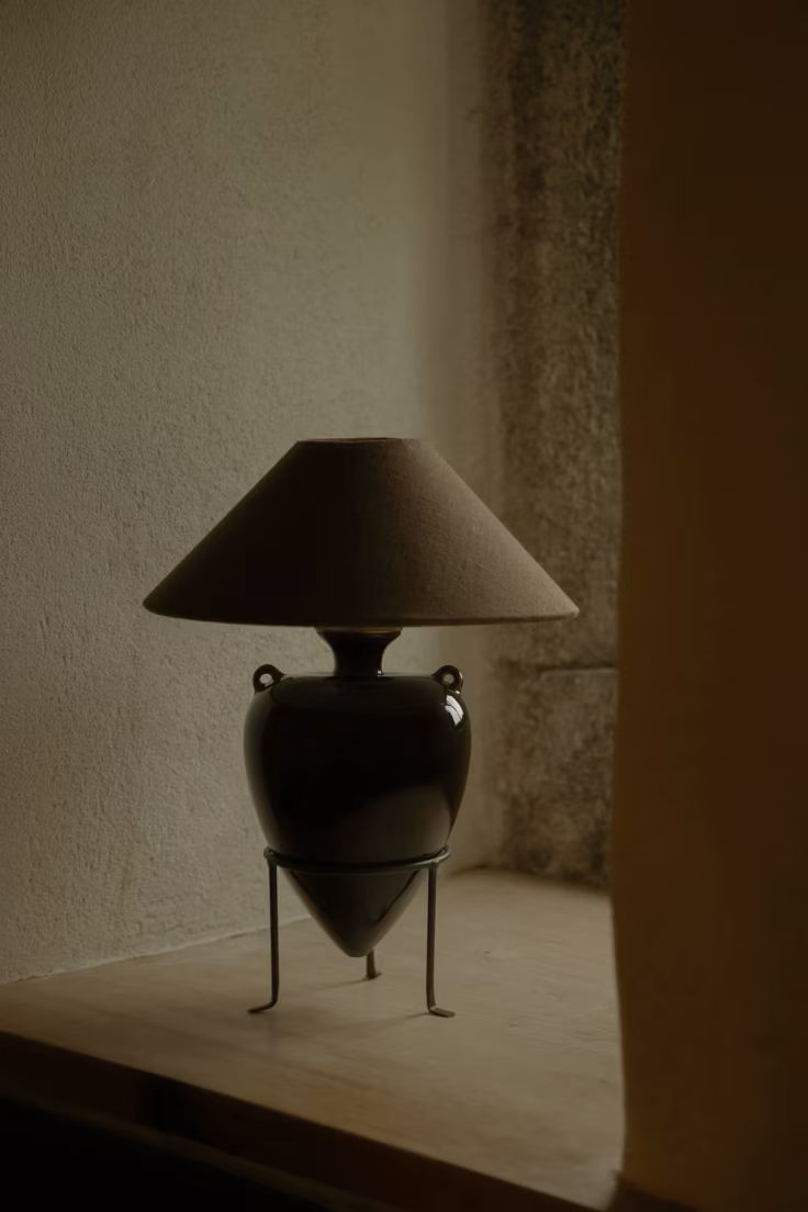 A decorative table lamp with a black ceramic base, tripod stand, and a beige lampshade, placed on a wooden surface against a textured wall.