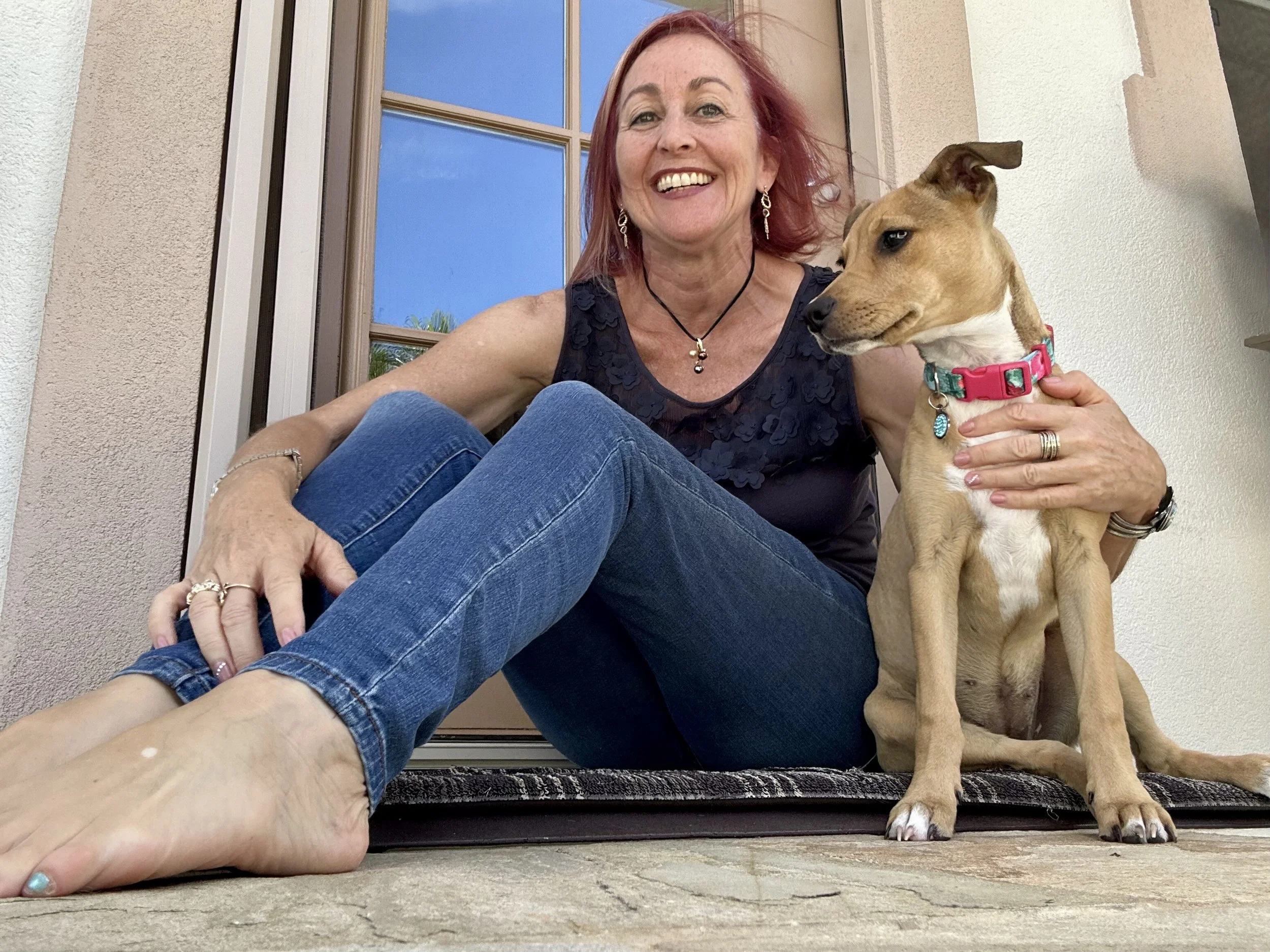 A smiling woman with red hair sitting on a porch with a tan dog wearing a pink collar.