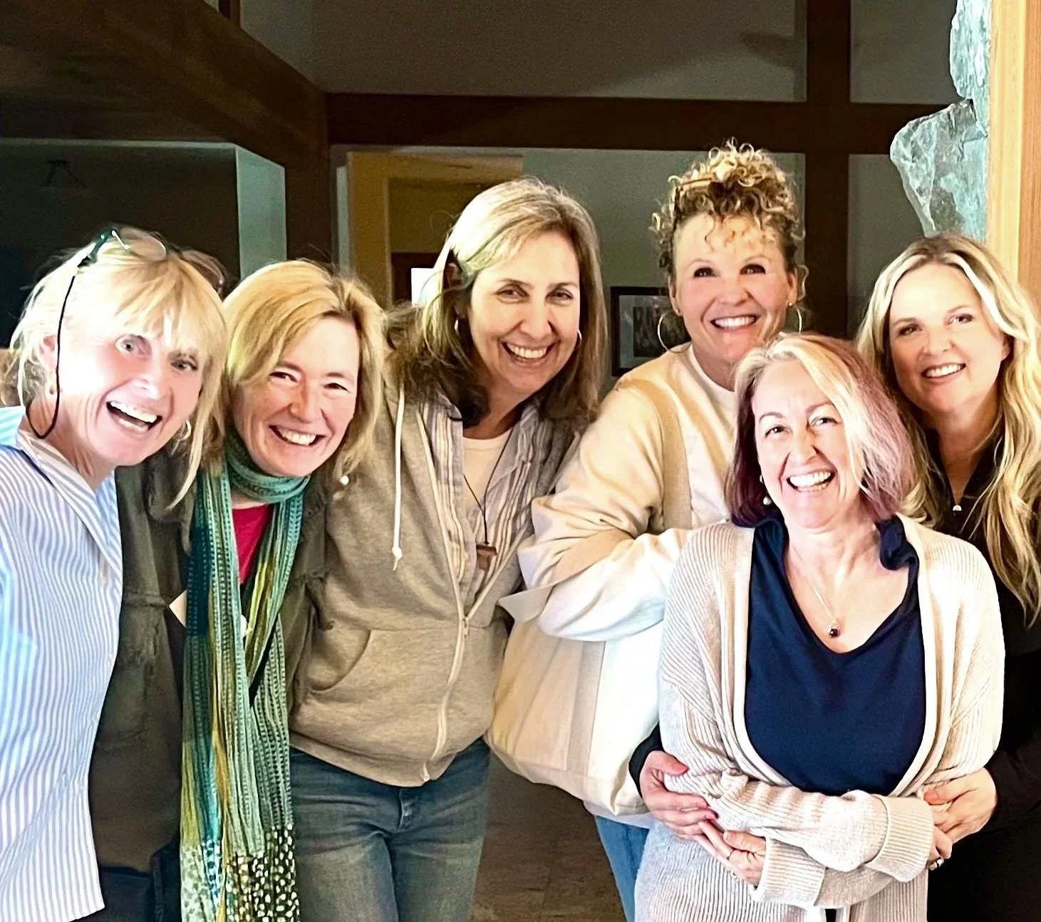Group of seven smiling women standing close together indoors, wearing casual clothing.