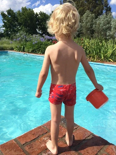 A young child with blonde hair in red swim trunks standing at the edge of a swimming pool, holding a red bucket, with greenery and bushes in the background.