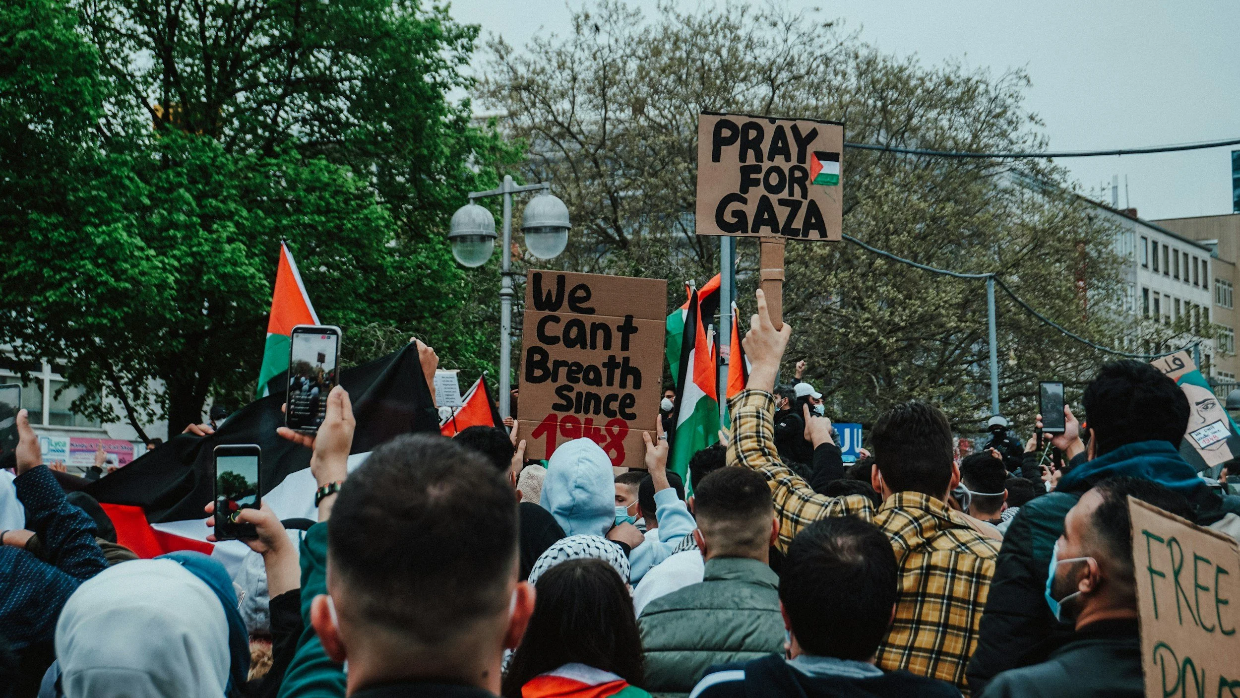 A large crowd of protesters gathered outdoors, holding signs with messages supporting Gaza and Palestine, some flags, and many people taking photos or recording using smartphones, with trees and city buildings in the background.