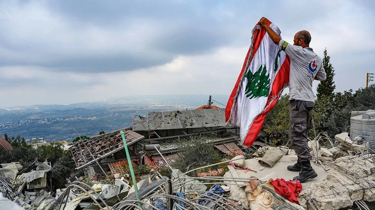 A man standing on debris and rubble, holding a Lebanese flag with a green cedar tree emblem, during a cloudy day with a distant landscape in the background.