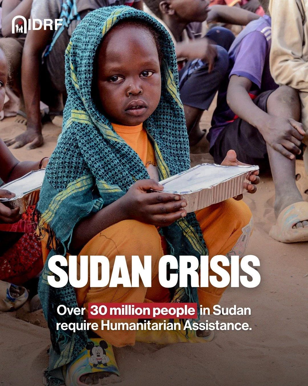 A young child with a scarf over their head sitting barefoot on the ground, holding a tray while surrounded by other children, during a humanitarian aid event in Sudan.