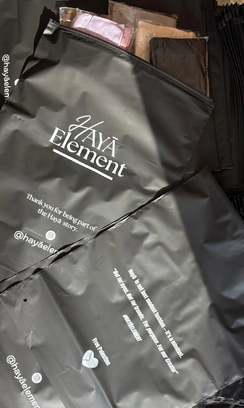 Black plastic shopping bags from 'Haya Element' with white text, containing various folded fabric items such as towels or linens. Some bags are partially open, showing the contents inside.