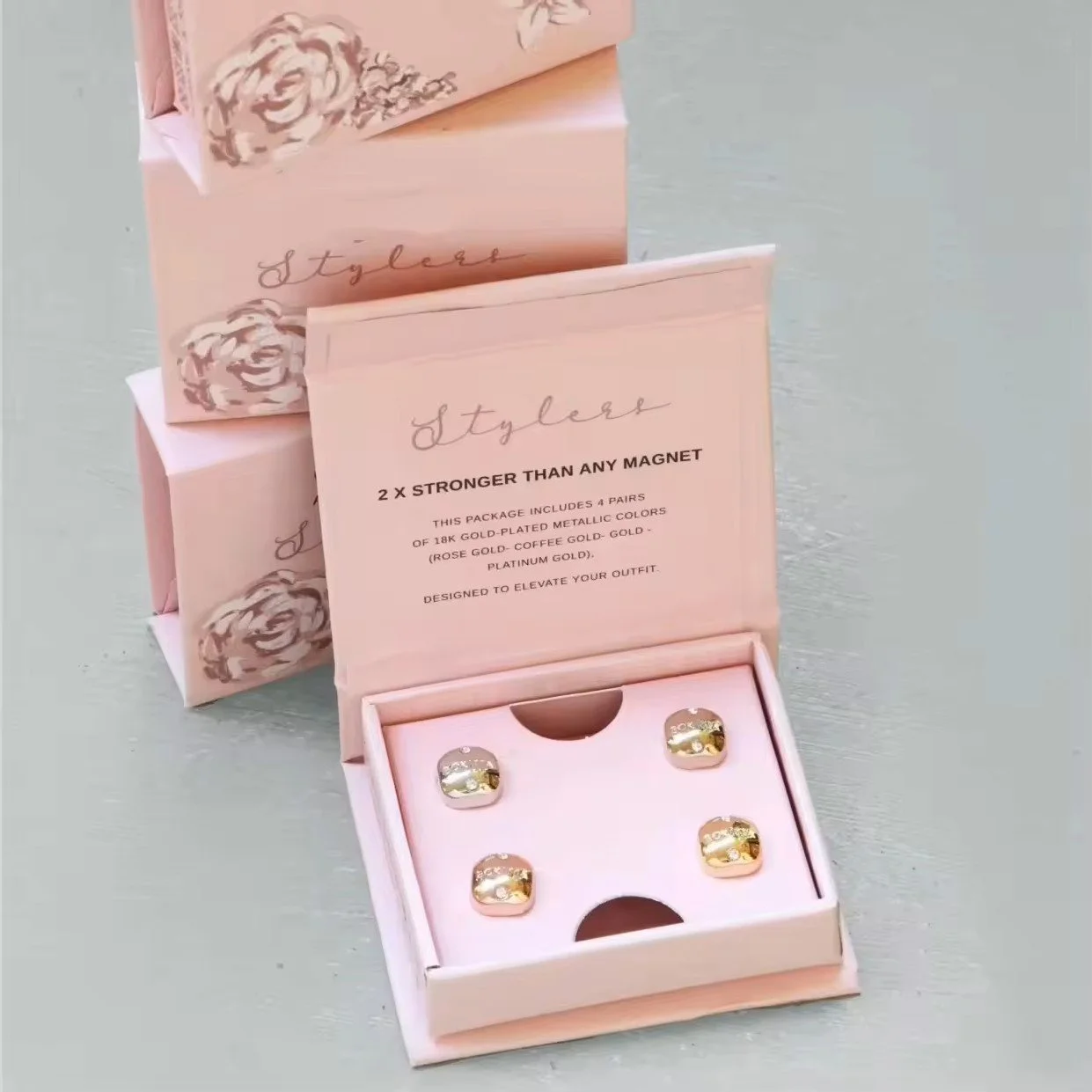 Pink jewelry box containing four pairs of gold-plated metallic earrings with words indicating they are stronger than magnets, displayed on a light-colored surface with packaging in the background.