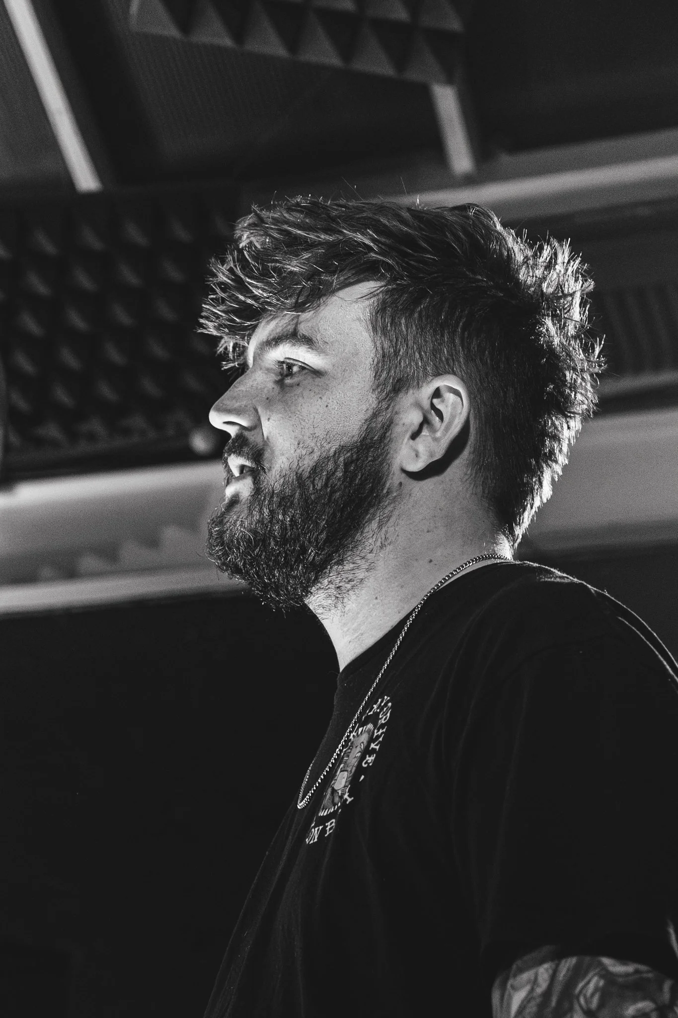 A black and white photograph of a young man with a beard and styled hair, looking to the left. He is wearing a dark t-shirt with a graphic on it and a chain necklace. The background contains soundproofing foam panels.