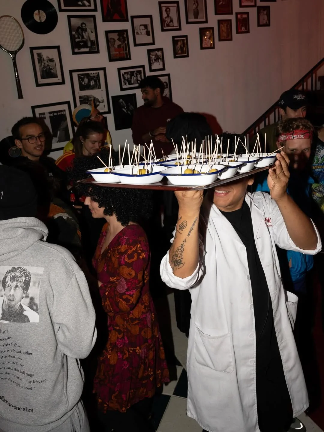 A chef carrying a tray with small bowls containing appetizers, each with toothpicks, in a crowded room with a wall decorated with framed photos and a tennis racket.