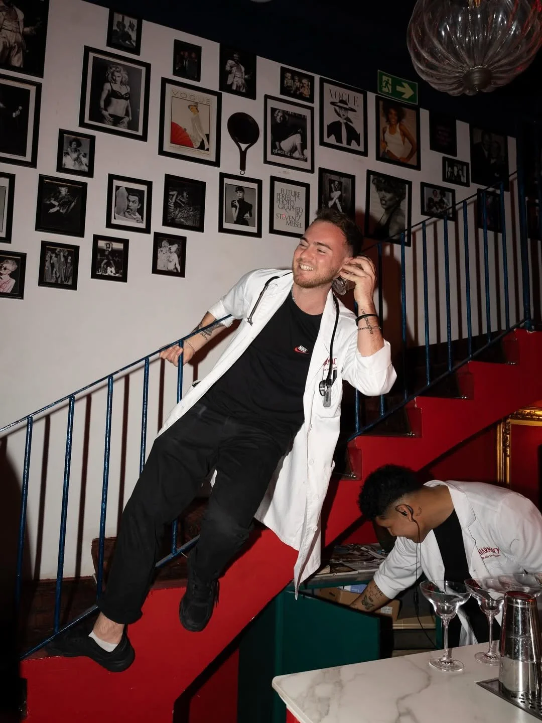 A man in a white doctor's coat, black shirt, and black pants leaning on a staircase railing, talking on a phone and smiling, with a person in a lab coat working below him at a table with glasses and barware, against a wall decorated with framed photos and magazine covers.