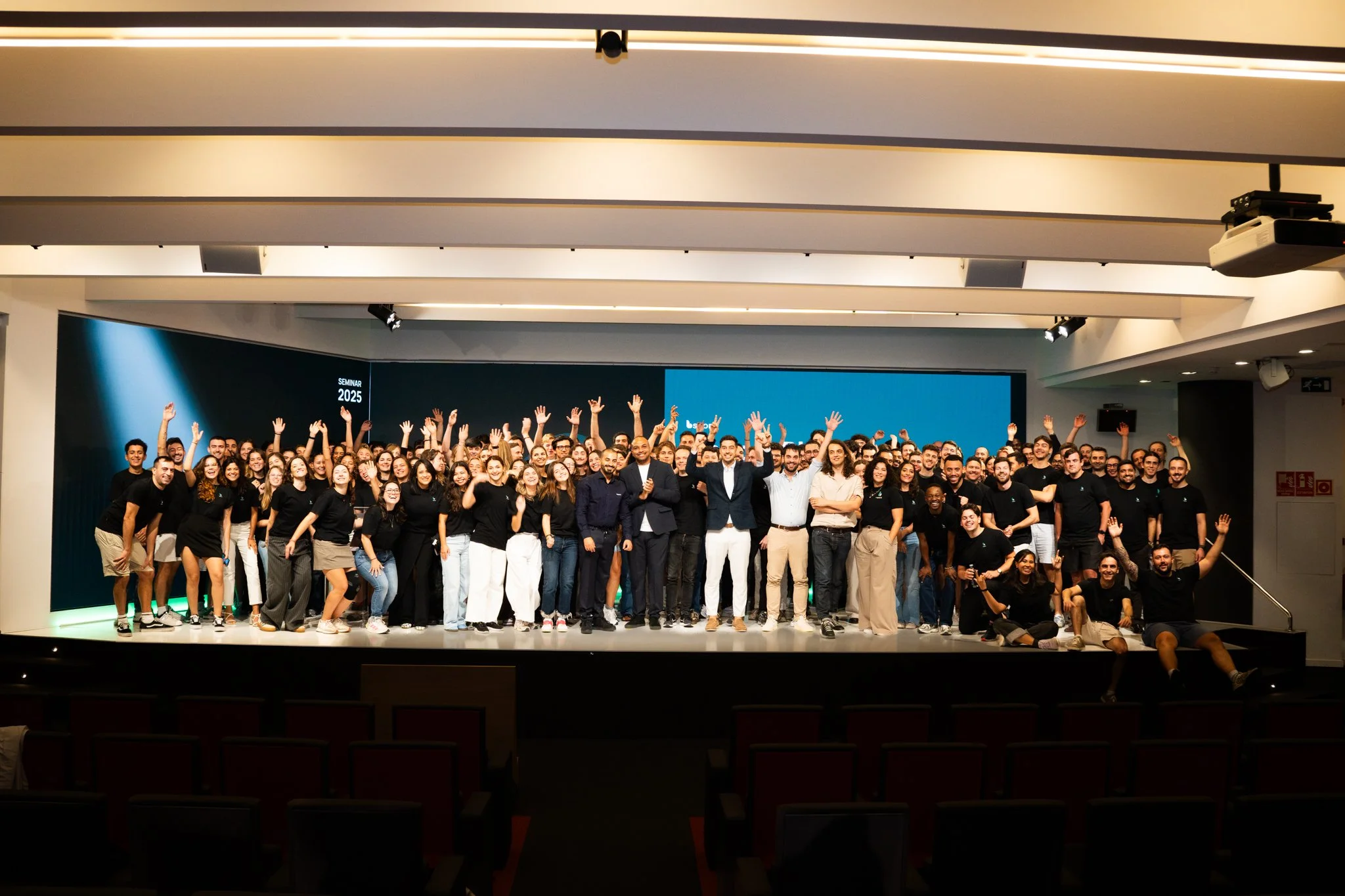 Large group of people on a stage in a conference room, smiling and waving, with a digital screen background showing 'SEMINAR 2025'.