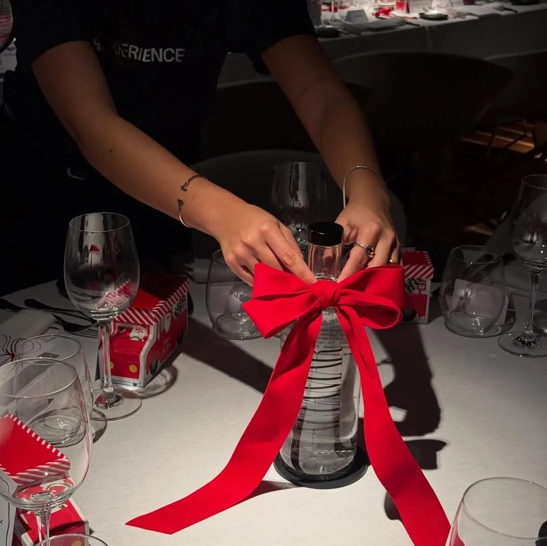 Person tying a red ribbon into a bow on a glass vase on a table set for a holiday celebration.