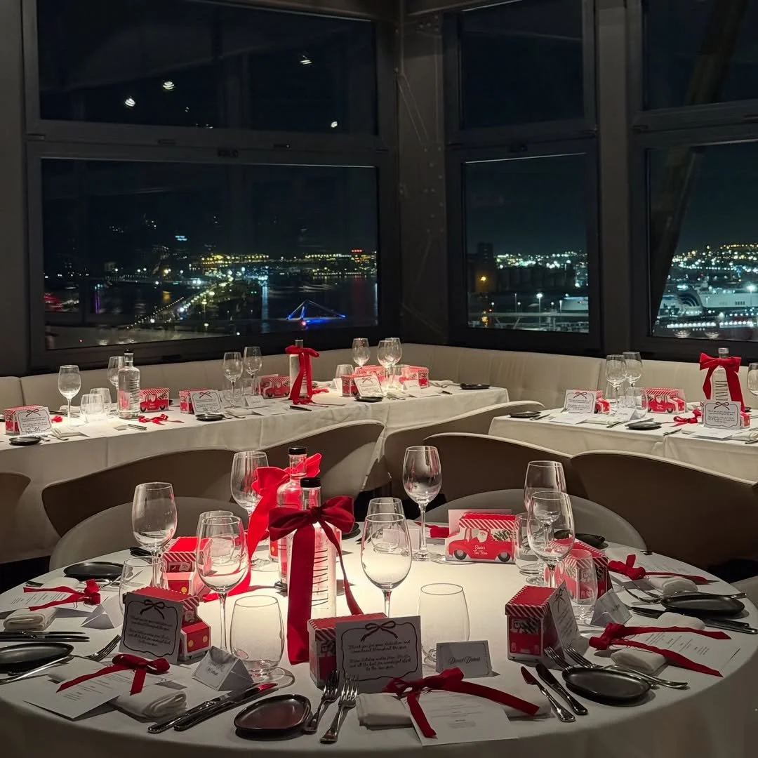 A restaurant or event space decorated for a festive occasion with tables set with wine glasses, silverware, white napkins with red ribbons, and red and white gift boxes, overlooking a cityscape at night through large windows.