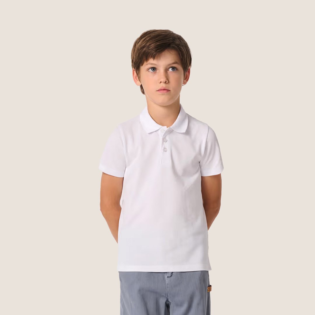 Jeune garçon souriant portant un polo blanc et un pantalon gris, debout contre un fond beige.