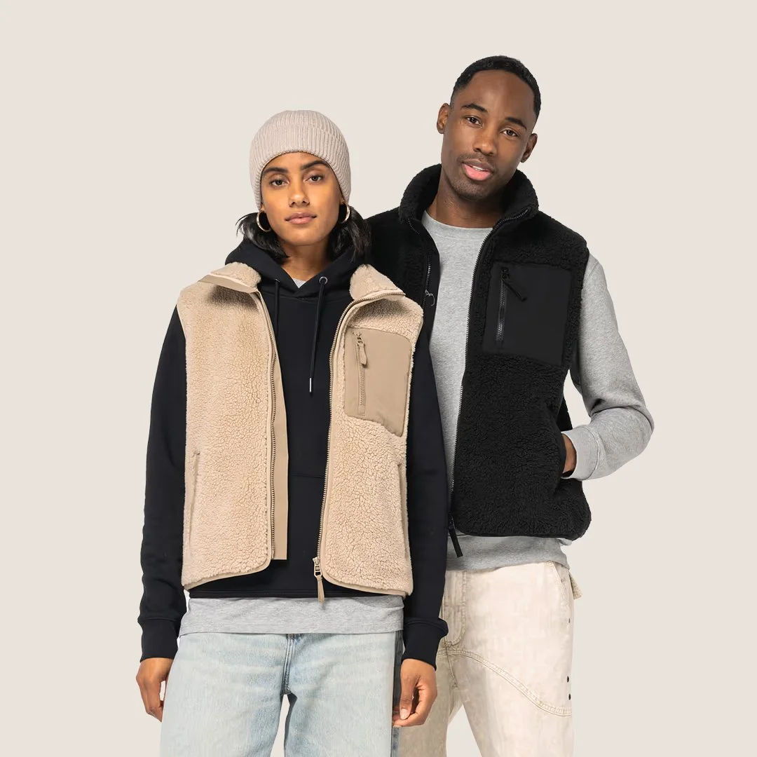 Deux personnes portant des vestes sans manches, la femme à gauche porte une veste beige en peluche, un sweat à capuche noir, un bonnet beige et un jean bleu clair, l'homme à droite porte une veste noire en peluche et un sweat gris clair, avec un pantalon beige.