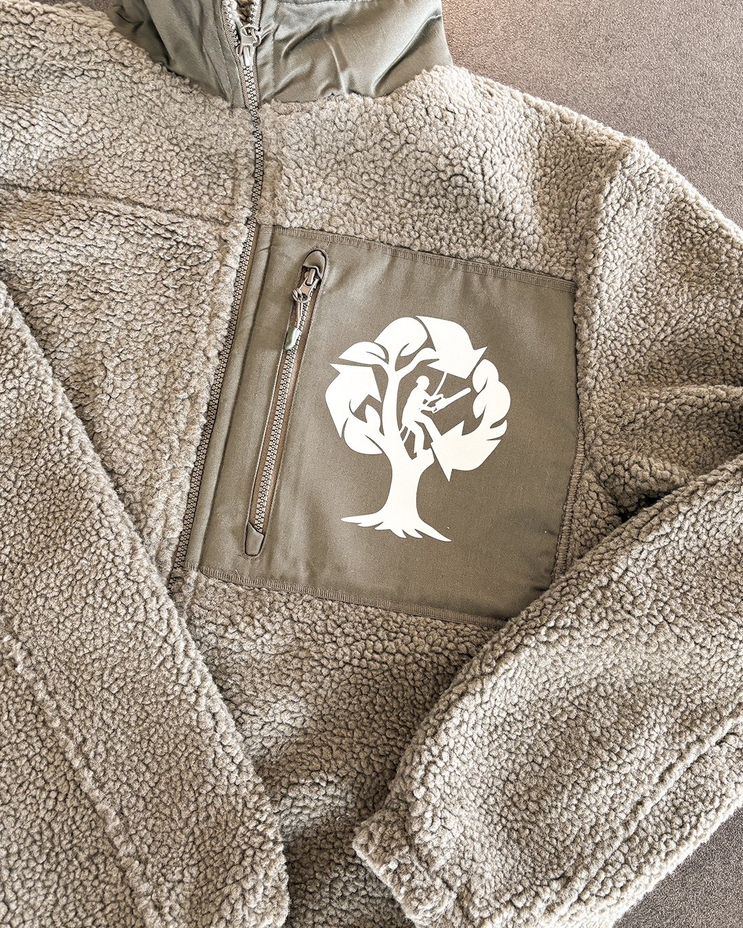 Veston en polaire beige avec poche en tissu olive et un logo représentant un arbre avec un escaladeur en son centre.