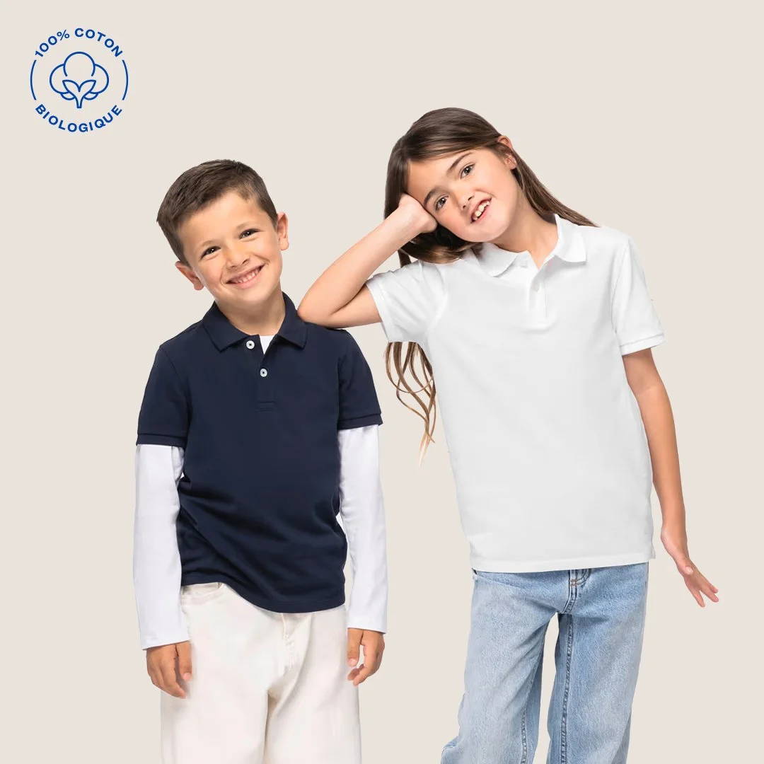 Deux enfants souriants, un garçon en polo bleu et une fille en t-shirt blanc, posant contre un fond neutre. Logo de coton biologique en haut à gauche.