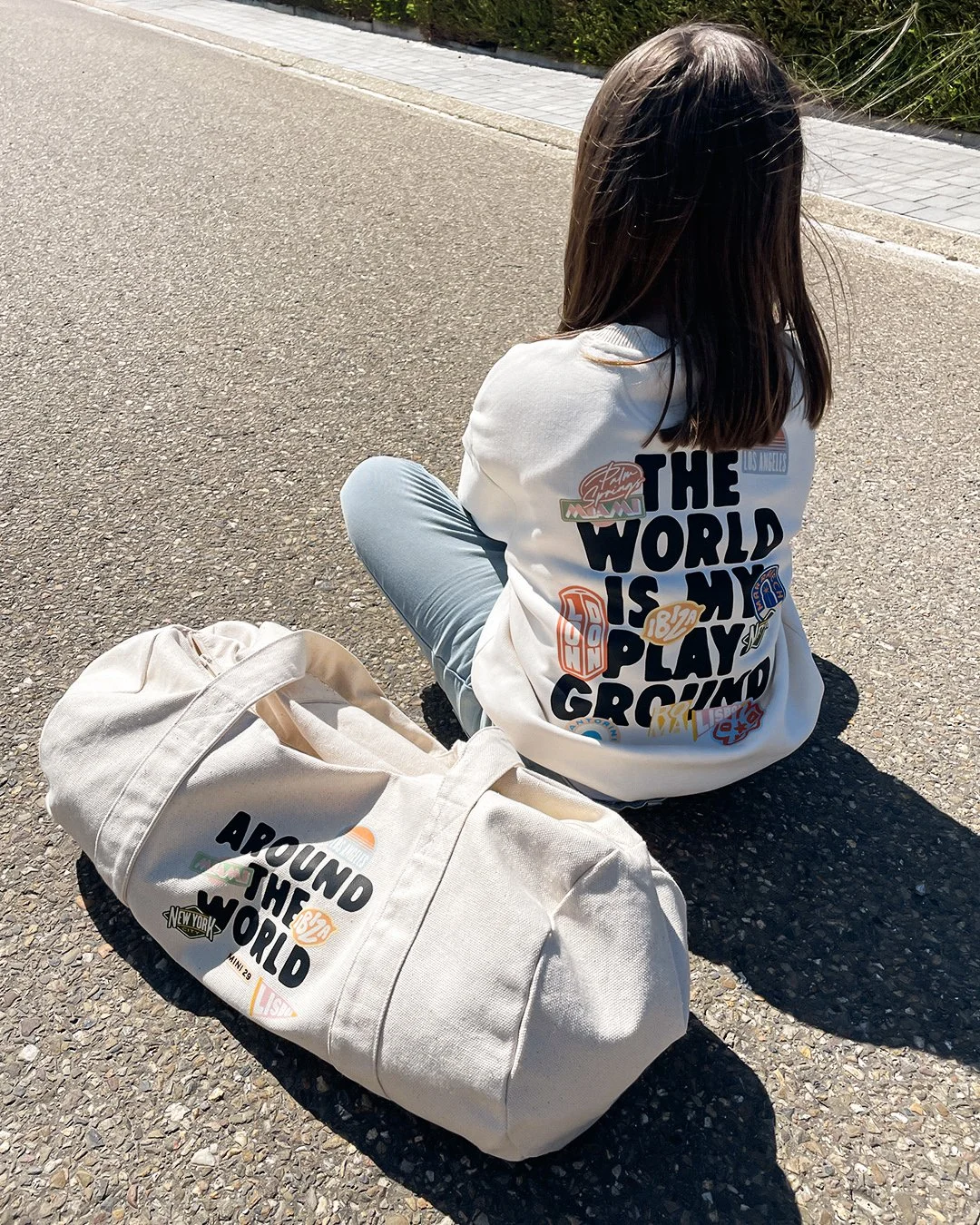 Jeune fille assise sur la route avec un sac beige à côté, portant un sweat-shirt blanc avec des affiches et textes en grands caractères.