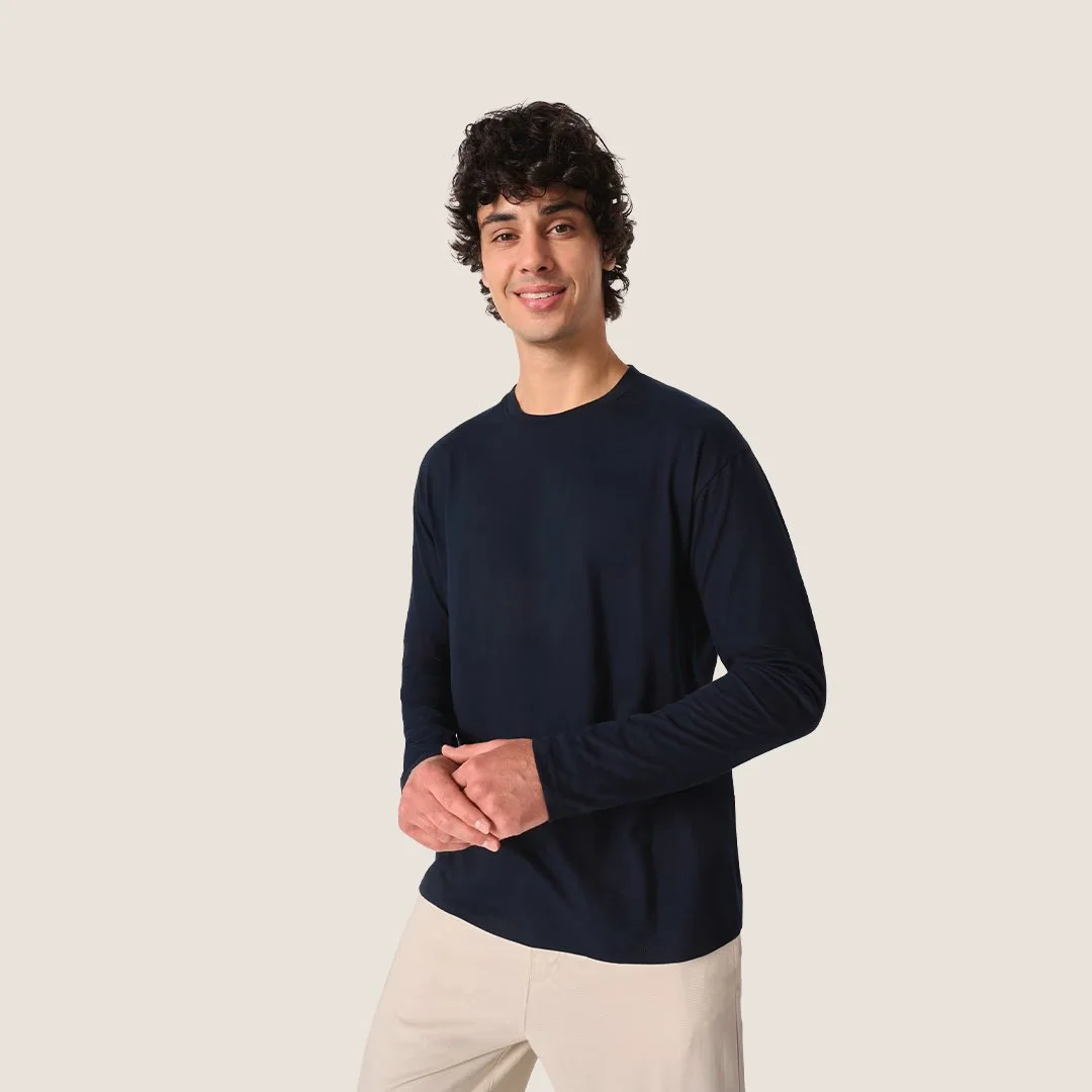 Jeune homme aux cheveux bouclés, portant un pull noir et un pantalon beige, souriant, posé contre un fond beige.