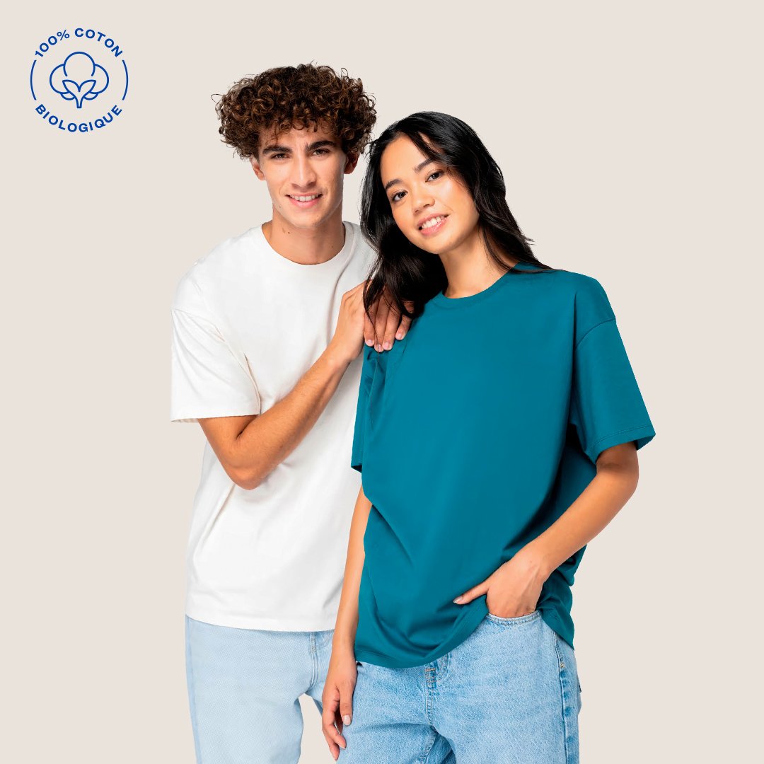 Deux jeunes adultes, un homme aux cheveux bouclés et une femme aux cheveux longs noirs, posent ensemble. Elle porte un t-shirt bleu et il porte un t-shirt blanc. Fond beige, logo en haut à gauche indiquant 100% coton biologique.
