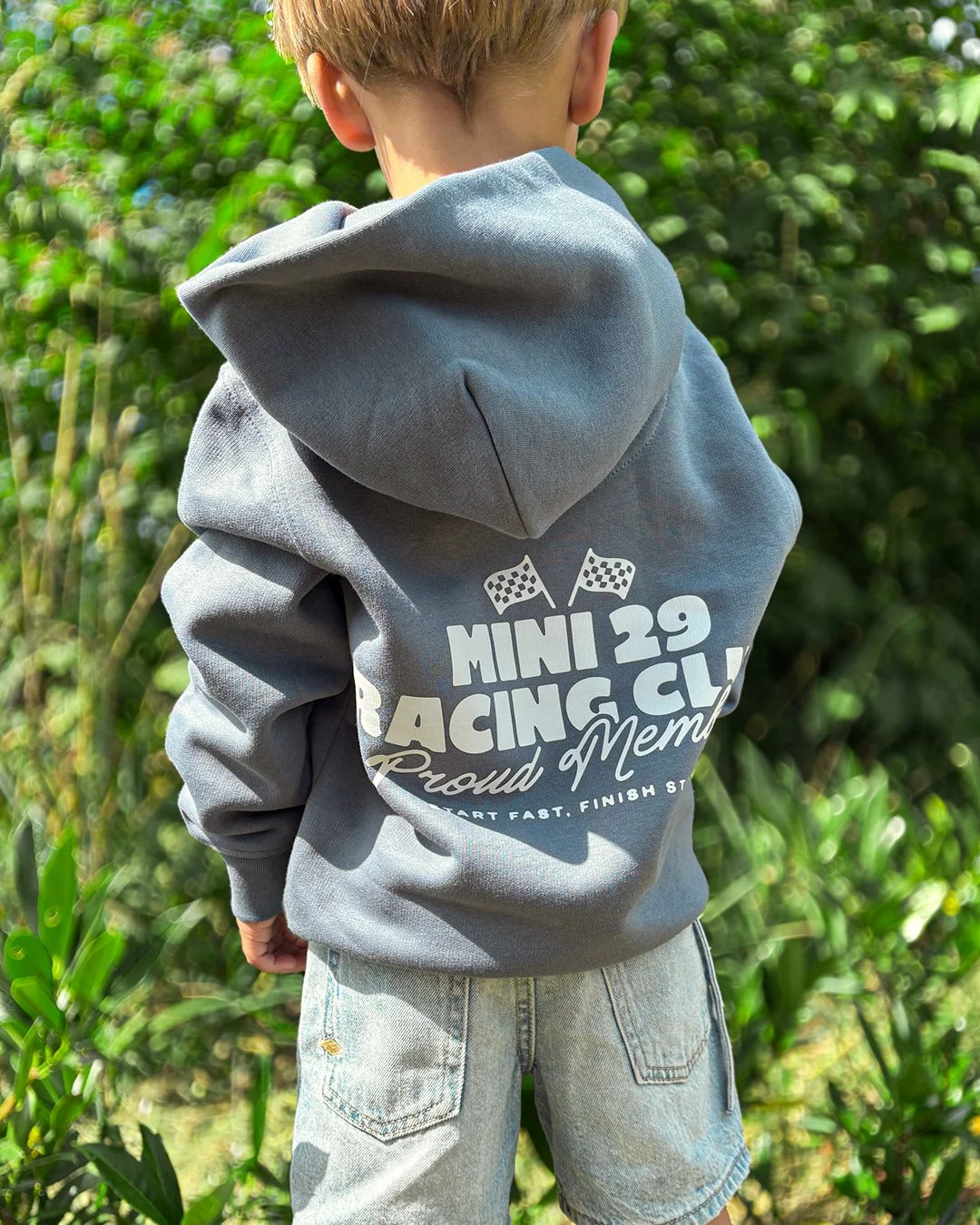 Enfant vu de dos portant un sweat à capuche gris avec un logo et la phrase 'Mini 29 Racing Club Proud Member' dans un environnement extérieur avec des plantes vertes.