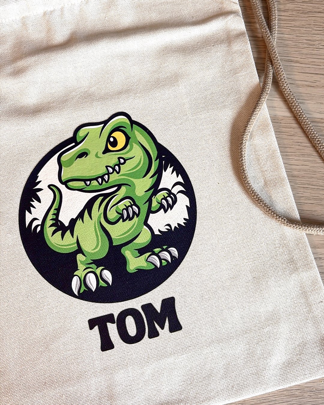 Sac en tissu beige avec un logo d'un dinosaure vert et le nom 'TOM' en dessous.
