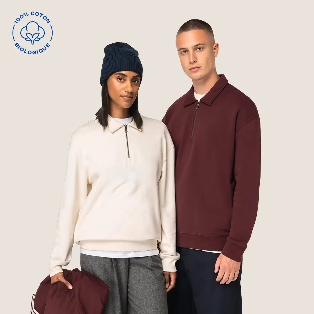 Deux jeunes adultes portent des vêtements en coton biologique, l'homme en sweat marron et la femme en pull beige avec un bonnet bleu, dans une image de mode minimaliste.