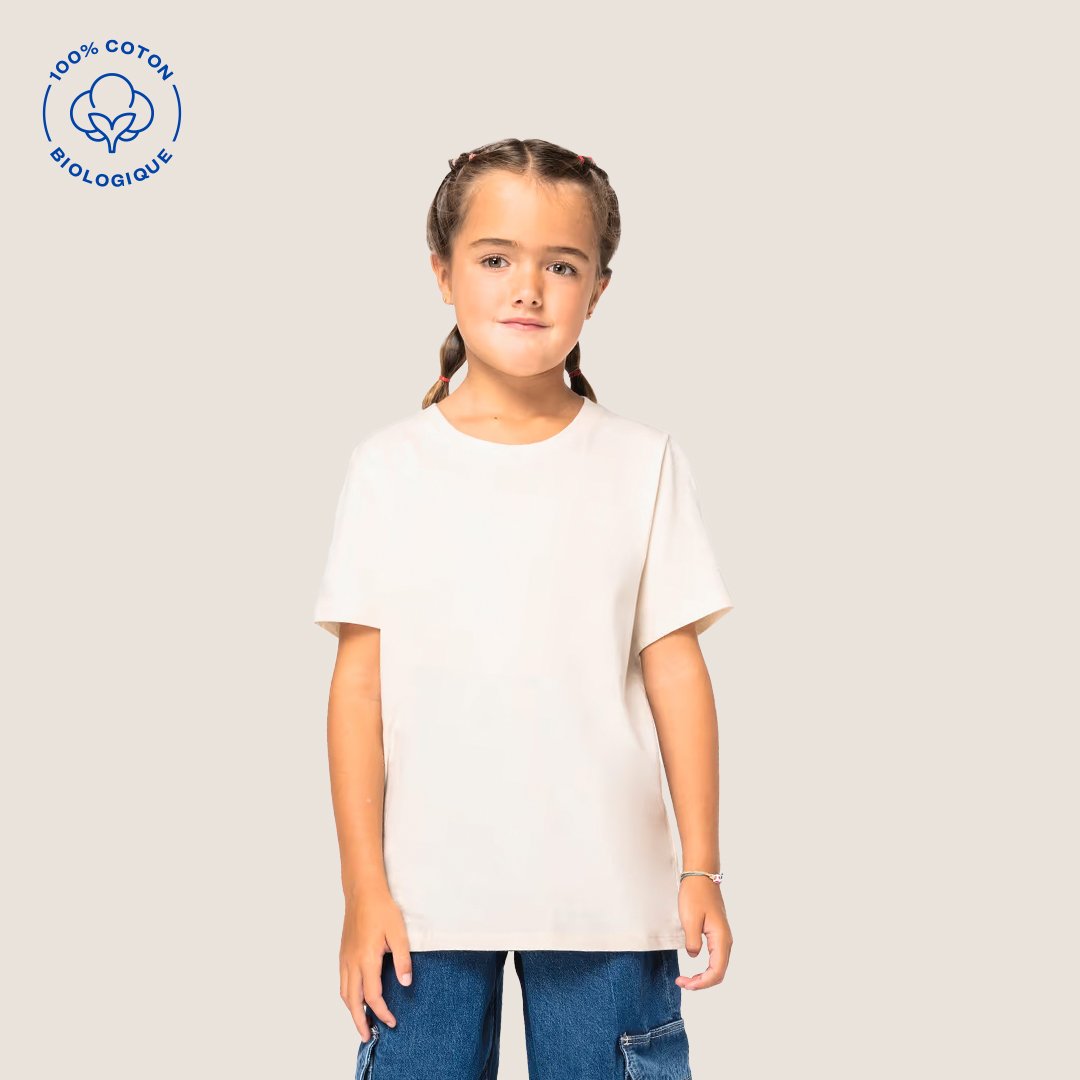 Jeune fille avec des cheveux bruns tressés, portant un t-shirt blanc et un pantalon en jean, debout devant un fond beige, avec un logo de coton biologique en haut à gauche.