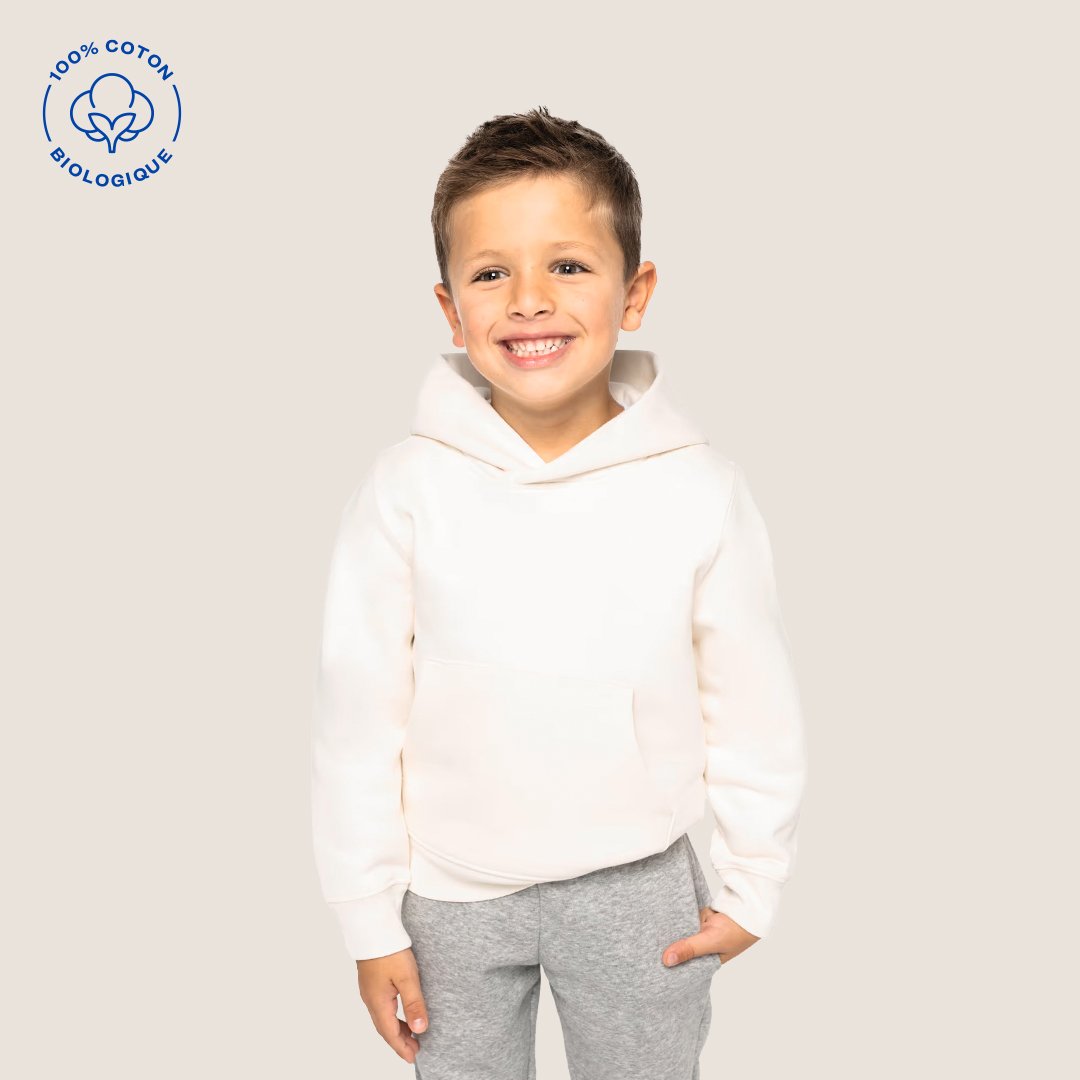 Jeune garçon souriant portant un sweat blanc et un pantalon gris, debout contre un fond beige avec un logo en haut à gauche indiquant coton biologique 100%.