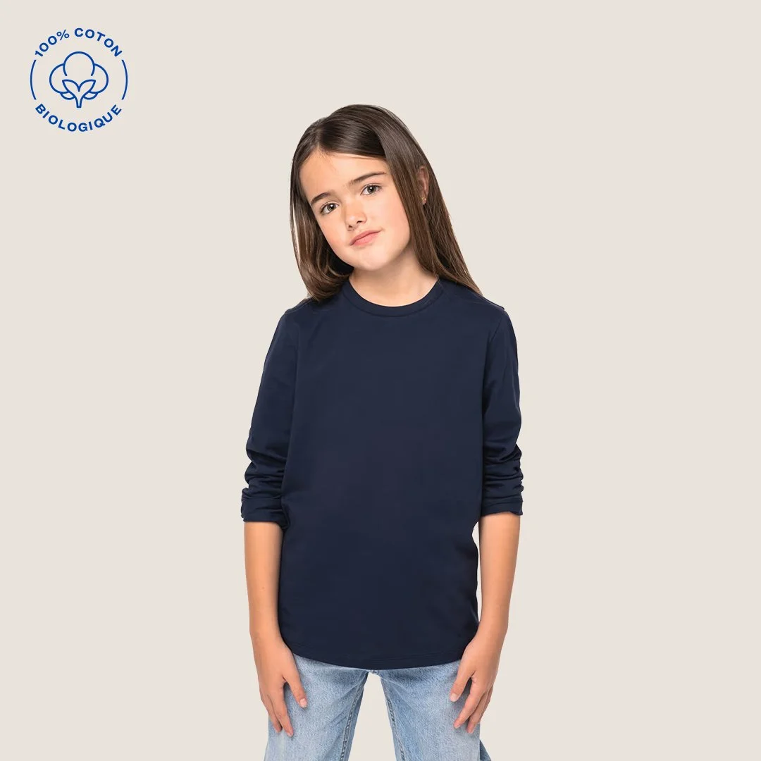 Jeune fille avec long cheveux bruns portant un t-shirt bleu marine et un jean clair, avec un fond neutre, logo de coton biologique à l'angle supérieur gauche.