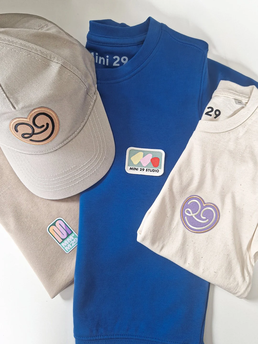 Vêtements et accessoires de la marque Mini 29, dont un chapeau beige avec un logo, deux t-shirts blancs et bleus avec des logos, et des autocollants »
