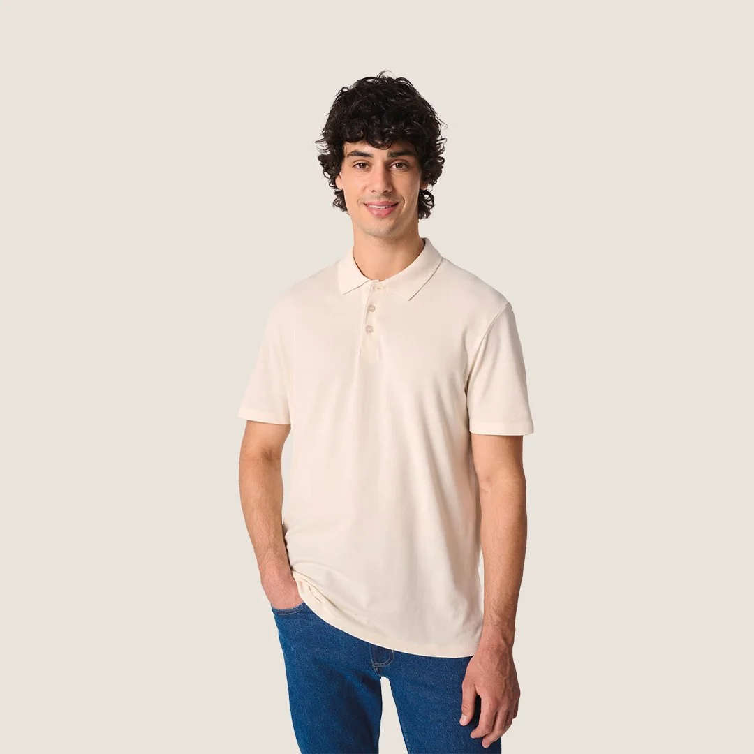Homme souriant avec cheveux bouclés, portant un polo beige et un jean bleu, debout contre un fond blanc.