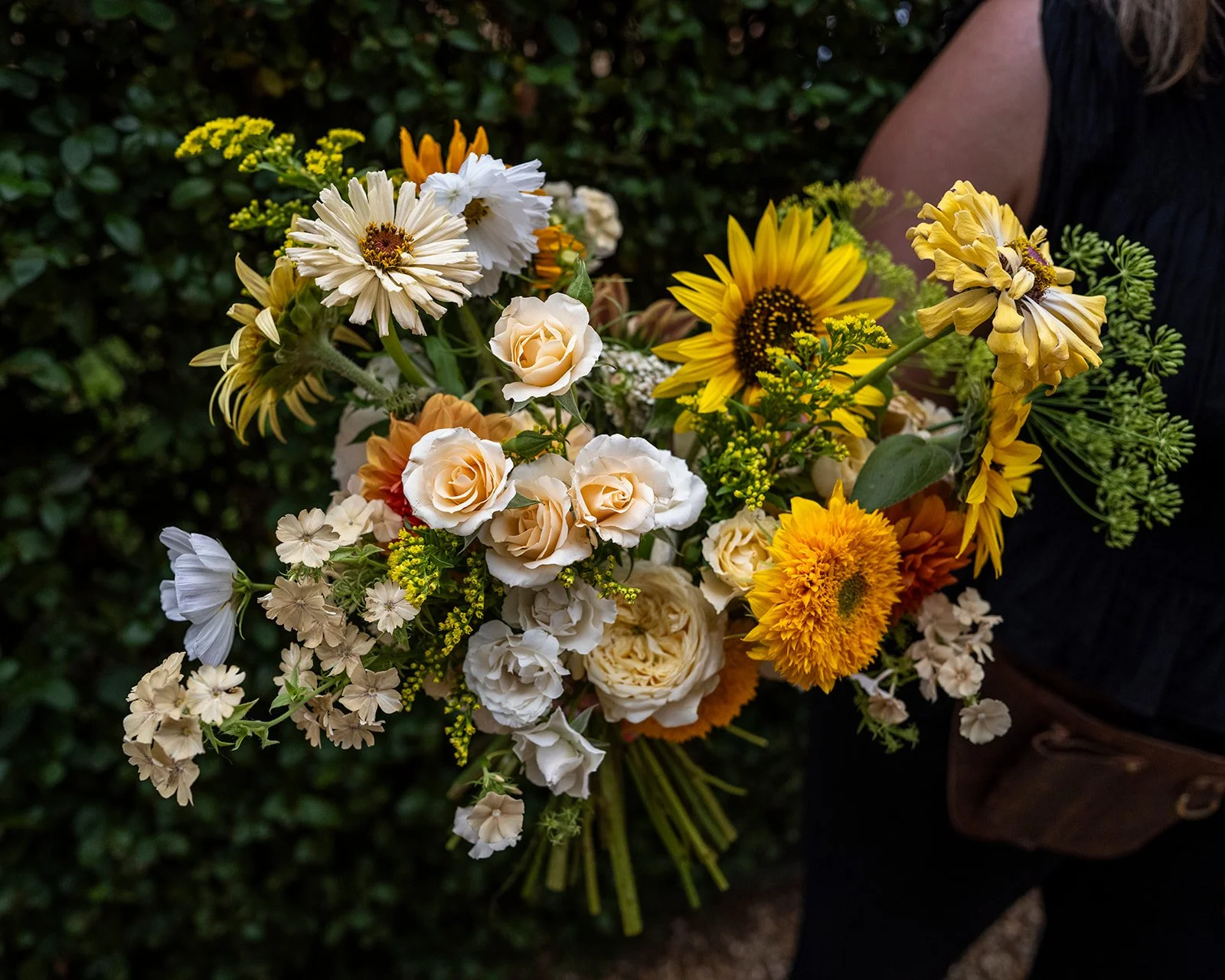 A wedding florist holds a wedding bouquet of yellow flowers including sunflowers, roses, and daisies.