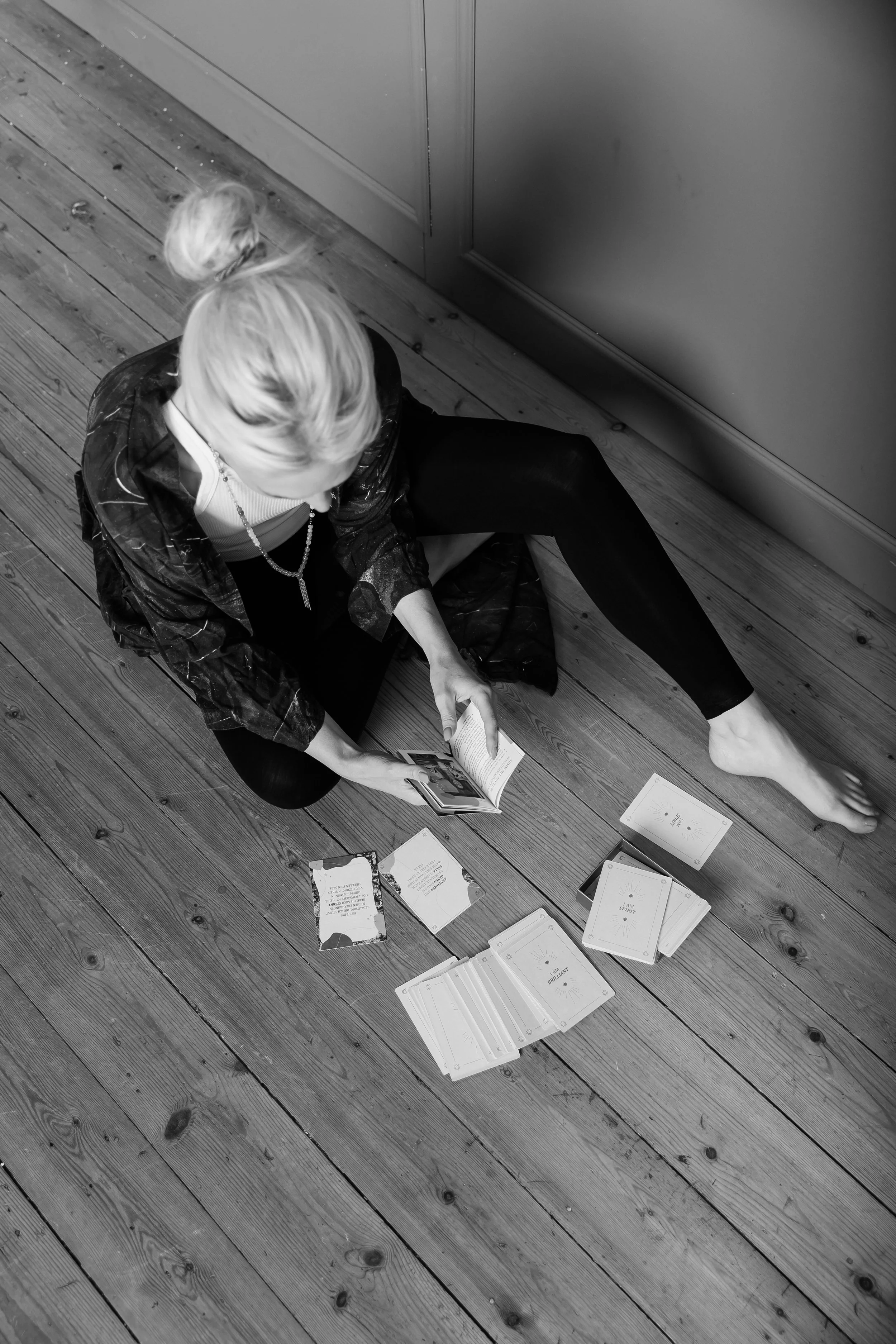 Eine Frau sitzt auf einem Holzboden und liest ein Buch. Vor ihr sind mehrere Karten und ein Notizbuch ausgelegt.