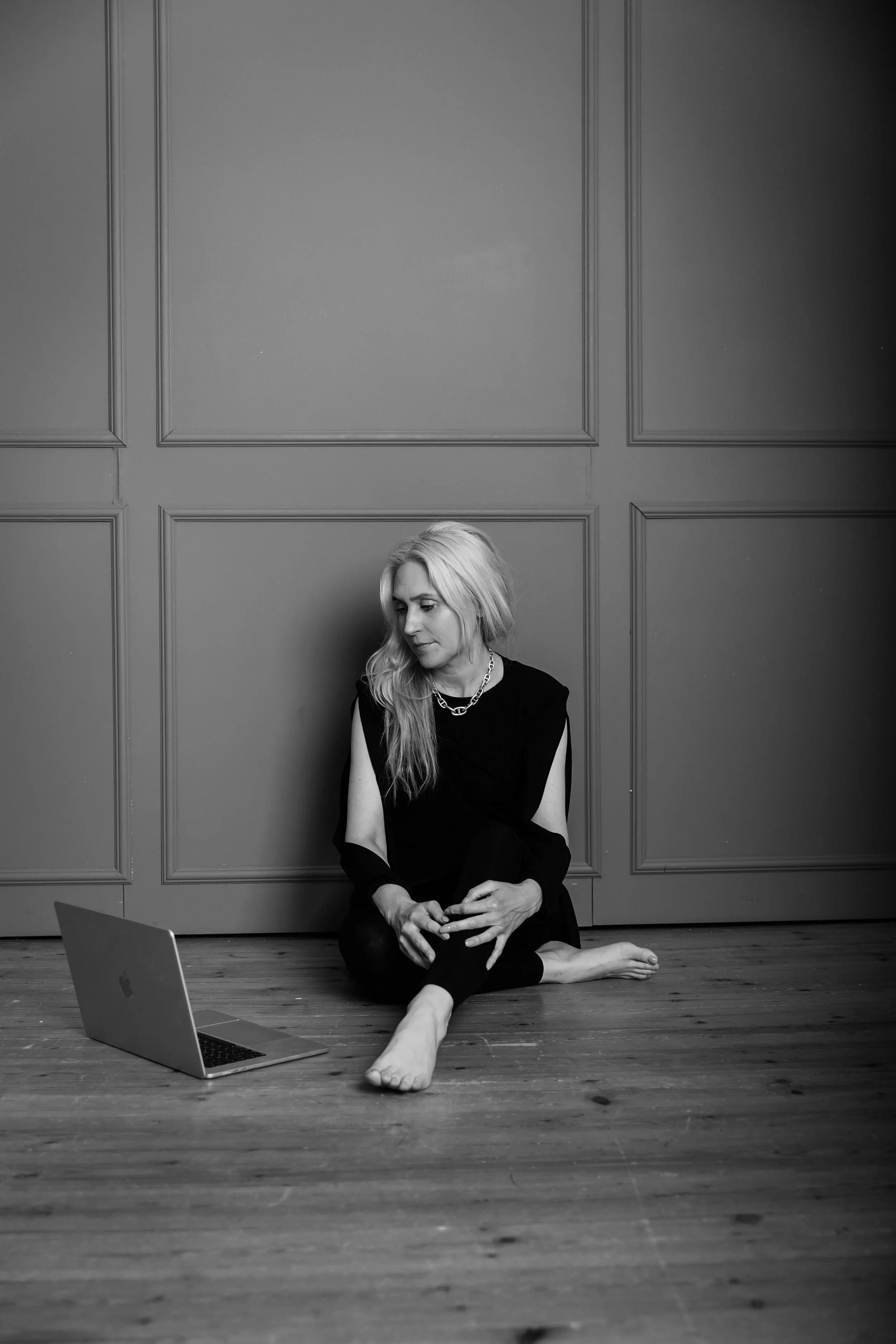 Eine Frau sitzt barfuß auf einem Holzboden vor einer Wand mit Paneelen. Sie trägt dunkle Kleidung, eine Halskette und hat lange blonde Haare. Vor ihr steht ein Laptop.
