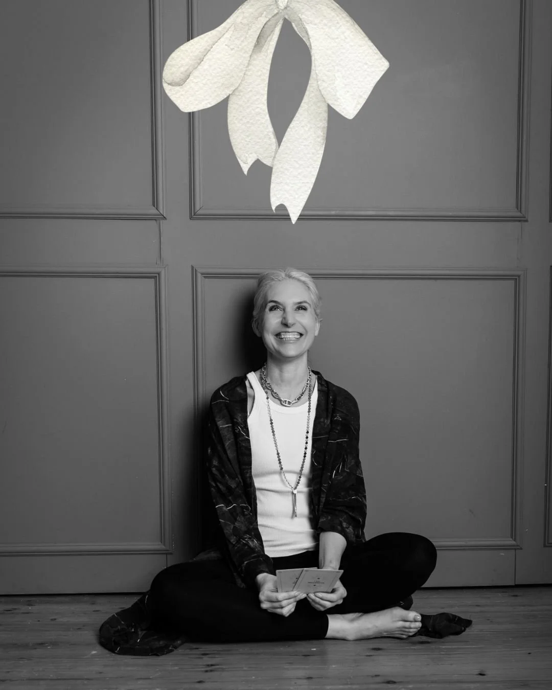 Eine lachende ältere Frau sitzt barfuß auf einem Holzboden vor einer Wand mit Verzierungen, hält Karten in den Händen, mit einem großen weißen Papierblumen-Dekor über ihr.