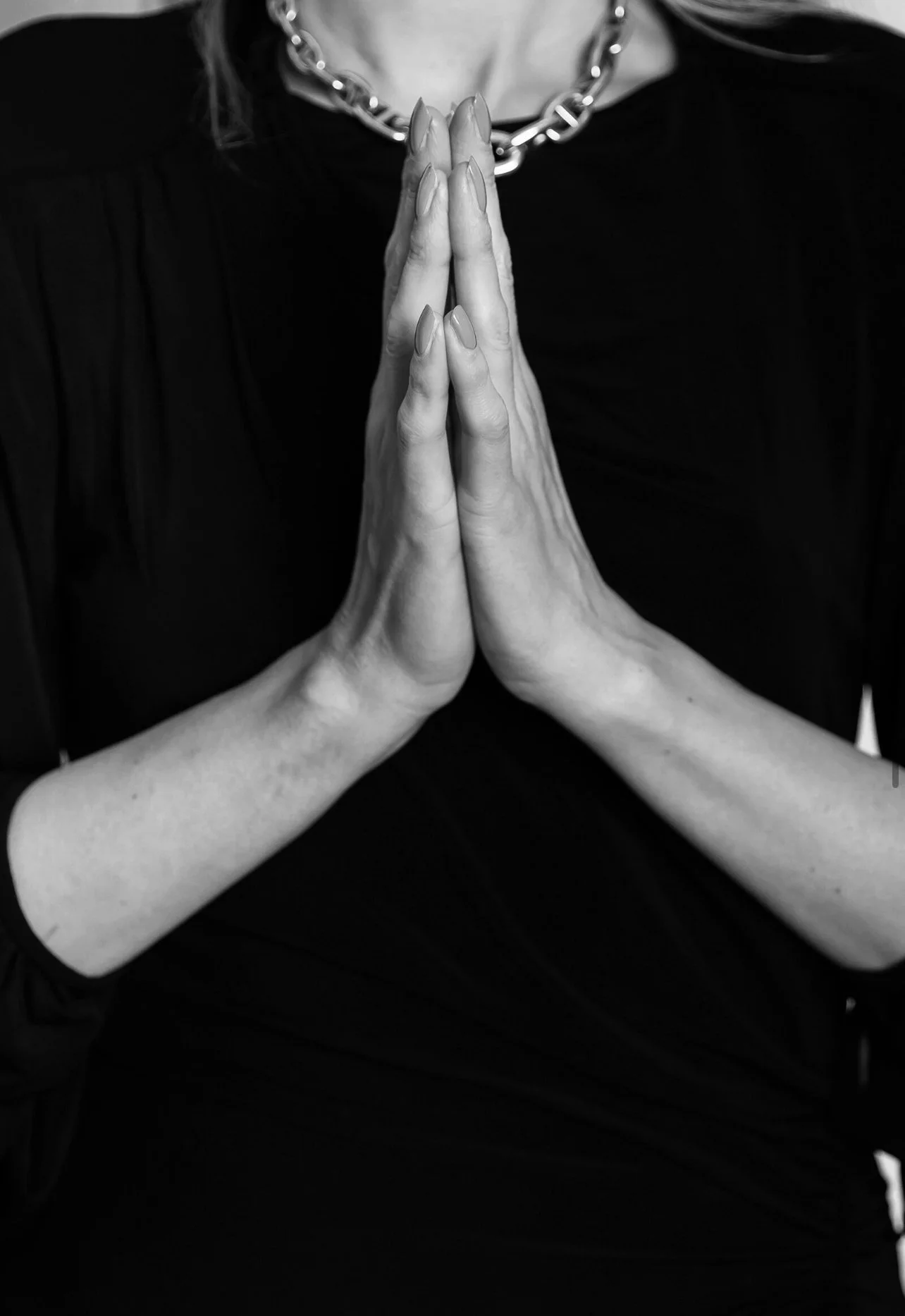Nahaufnahme von zwei Händen, die in Gebetshaltung zusammengelegt sind, vor einem schwarzen Hintergrund, eine Person trägt eine große Kette um den Hals.