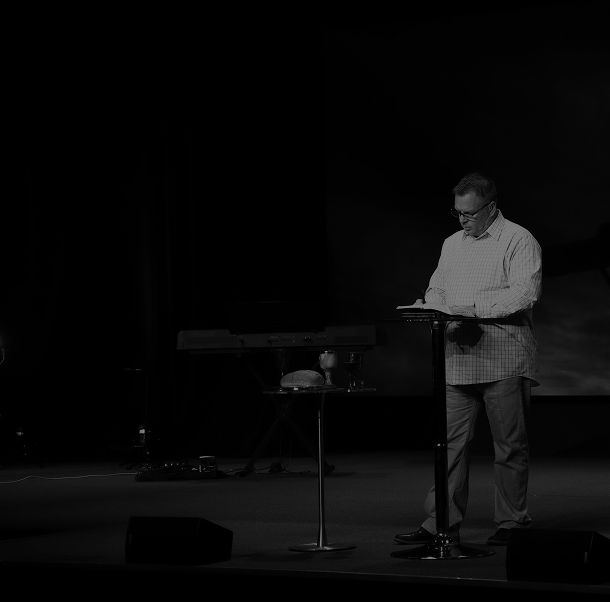 A man stands on a stage reading or speaking from a book or paper, with a table and a keyboard behind him, in a dark setting.