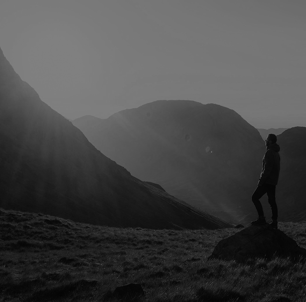 Person standing on a rock in a mountainous landscape, with mountains in the background and sunlight shining from the upper left.