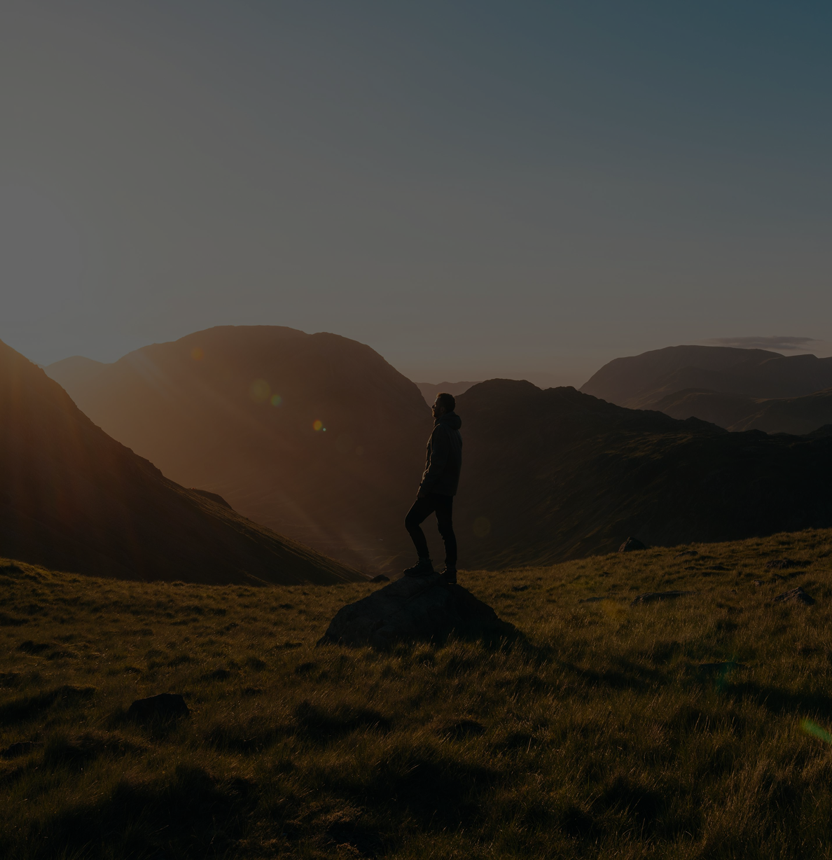 A person standing on a large rock in a mountainous landscape during sunset or sunrise, with the sun low in the sky casting warm light.