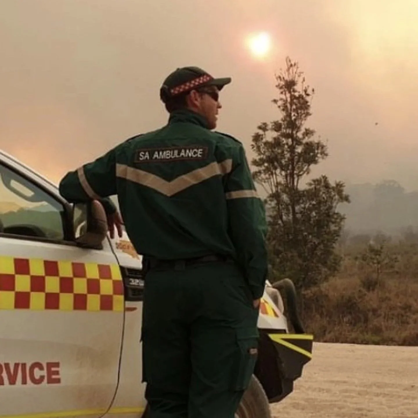 A person in a green SA Ambulance uniform stands beside an ambulance vehicle with a checkered pattern, during a smoky, hazy day with a sun and dry landscape in the background.