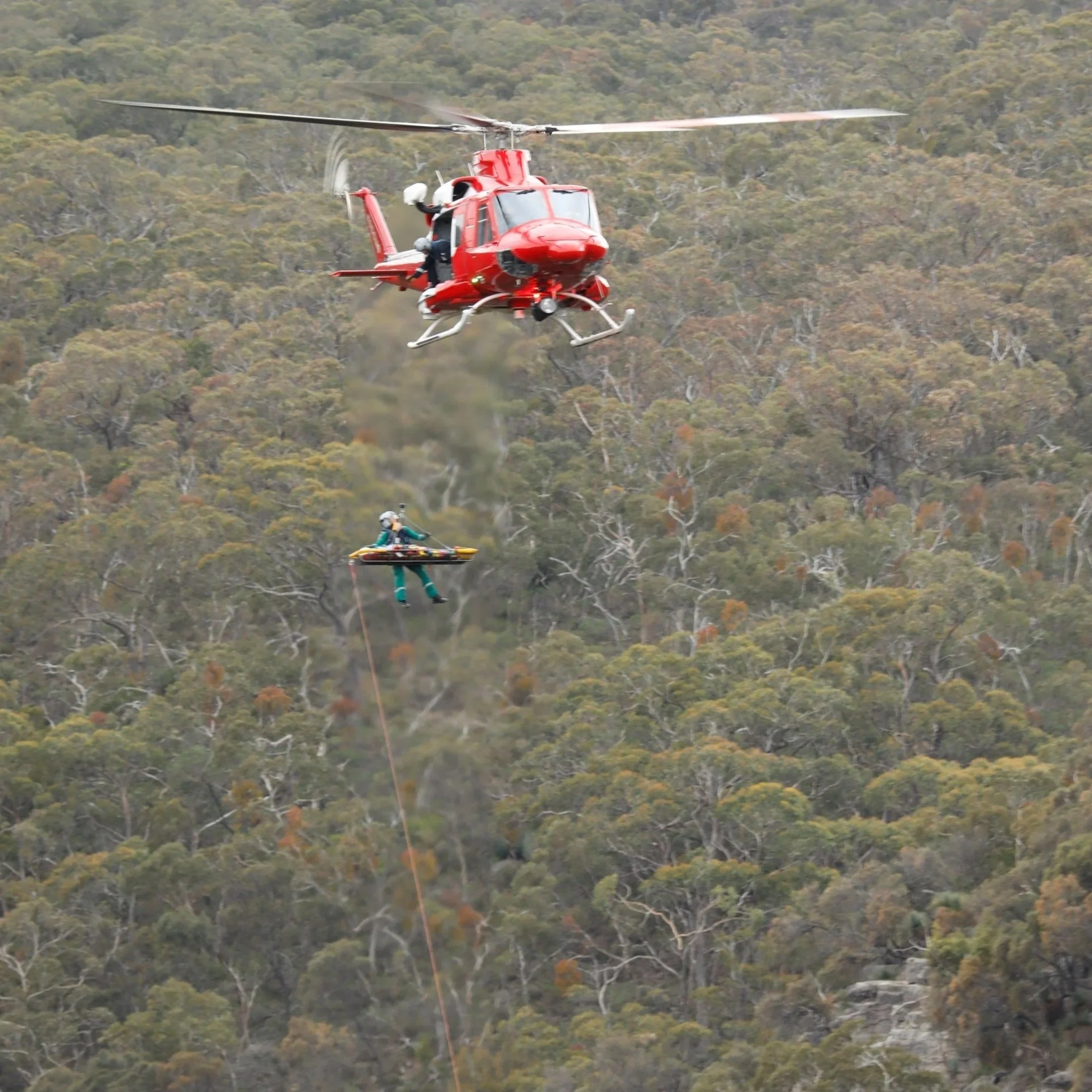 A red helicopter is flying over a forested area with trees in fall colors. A person is parachuting from the helicopter, descending toward the ground.