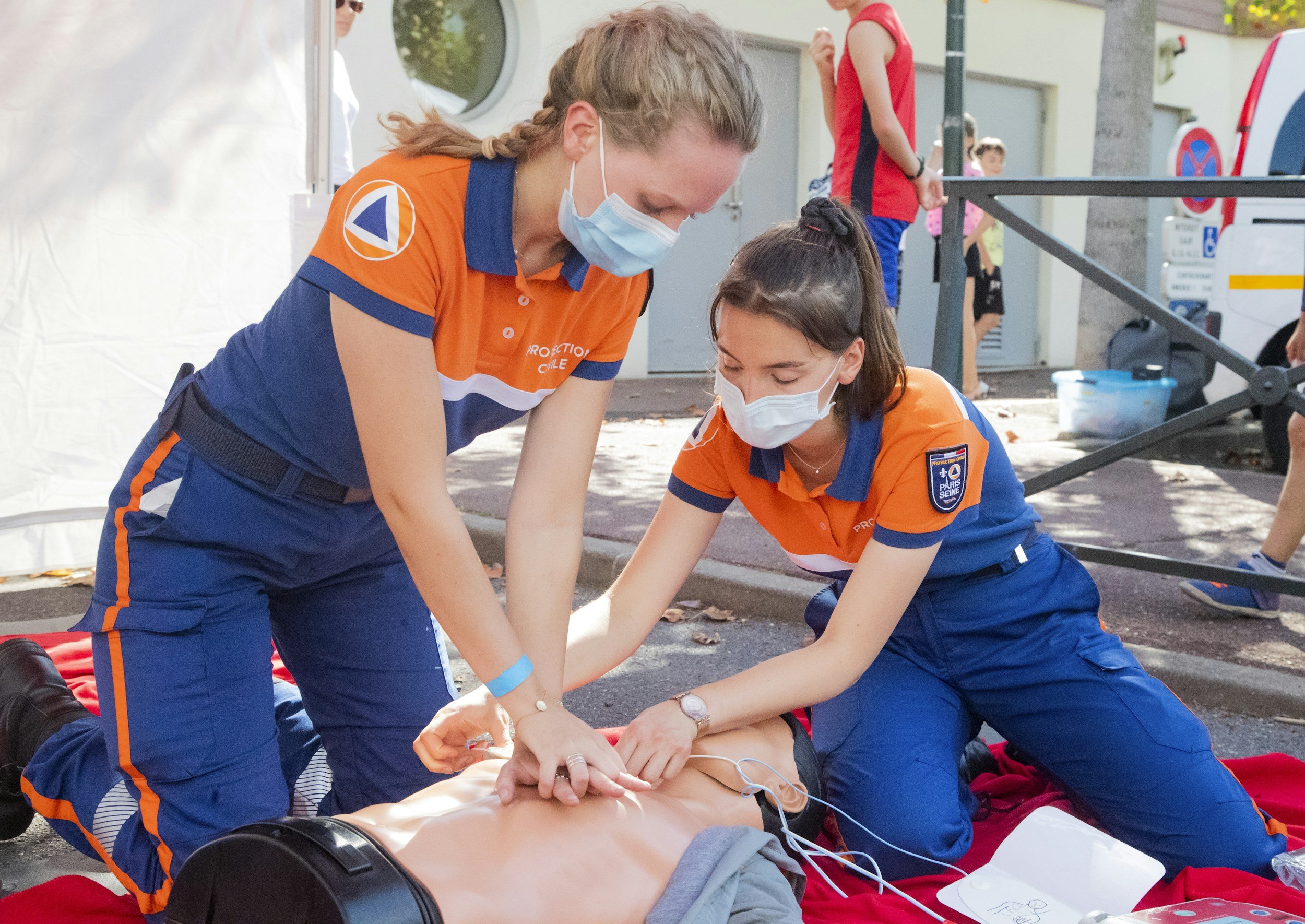 Two emergency medical responders in orange uniforms and face masks are providing CPR and medical assistance to a person lying on the ground, with other people and emergency vehicles in the background.
