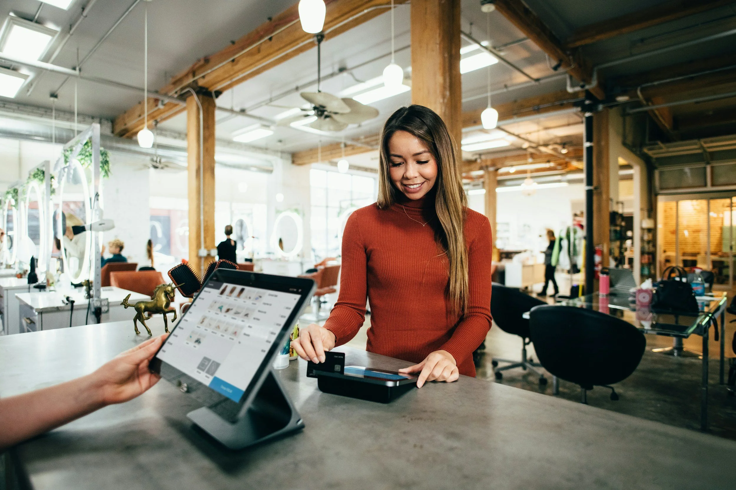 Woman with long brown hair wearing a red long-sleeved shirt working at a checkout counter in a decorated store, with a tablet and a small gold unicorn figurine on the counter, in a bright space with wooden beams and large windows.