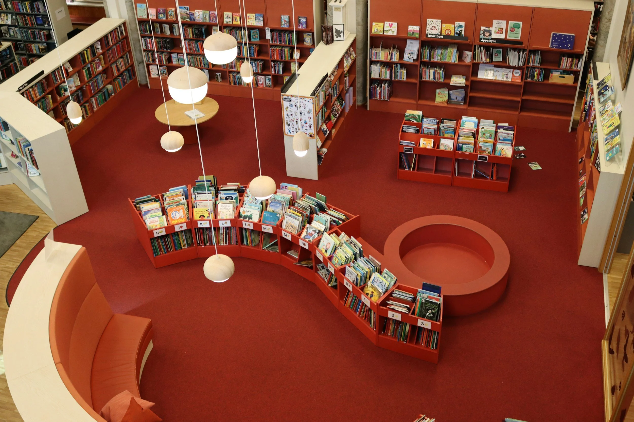 A children's section of a library with red bookshelves filled with children's books, a circular book display with more books, and a curved seating area with orange cushions. There are hanging pendant lights and a poster on the bookshelf.