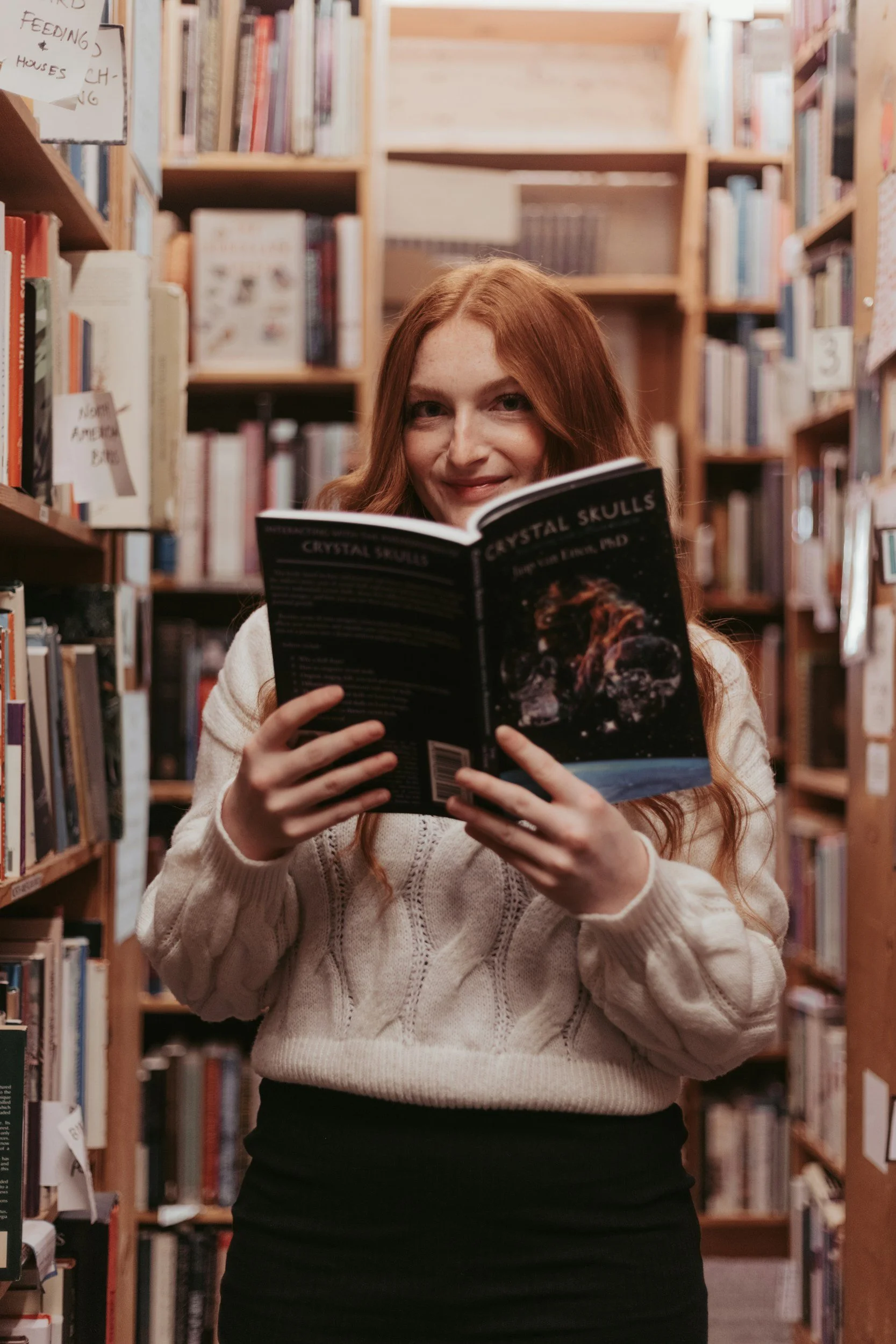 A woman with long red hair is smiling and reading a book titled 'Crystall Skulls' in a library surrounded by shelves filled with books.