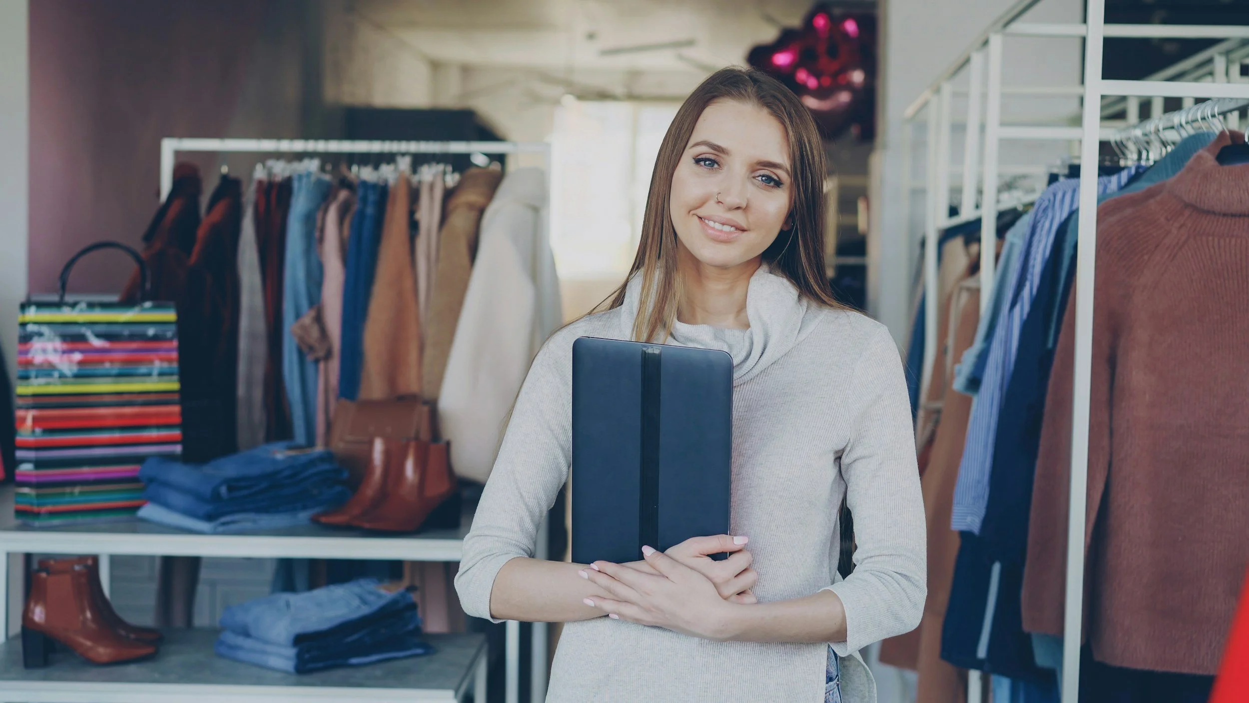A woman with long brown hair, blue eyes, and a nose piercing stands inside a clothing store, holding a black notebook. She is wearing a light gray sweater and smiling at the camera, with clothing racks and folded clothes around her.