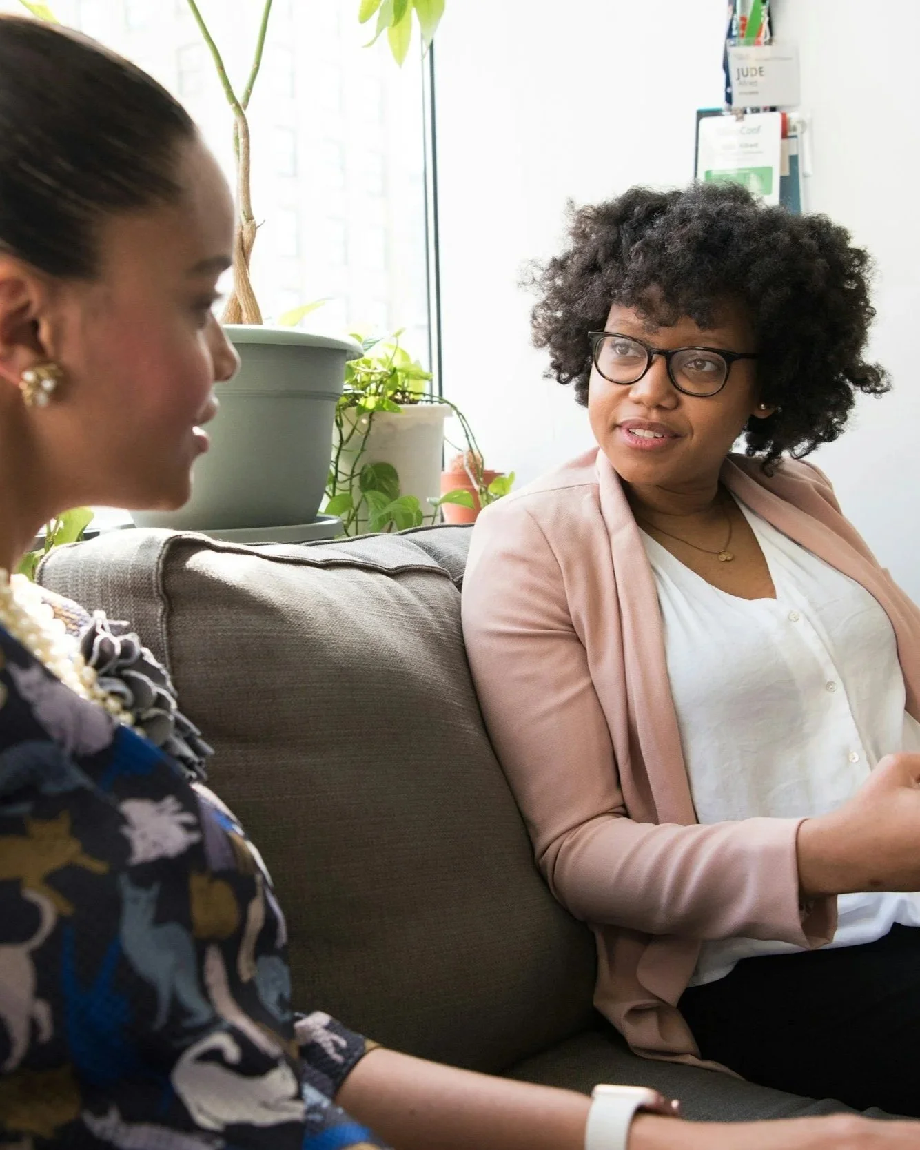 Two women are sitting on a gray couch in a bright room with large windows and green plants. They are engaged in conversation, with one woman on the left side of the image facing partially away and the woman on the right facing forward with glasses and curly hair.