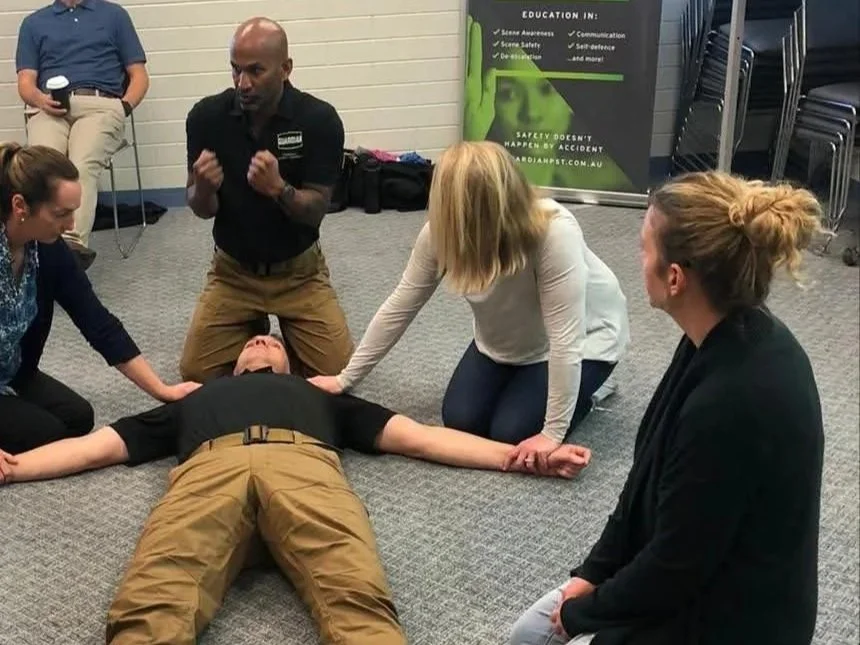 A group of people participating in a CPR training session indoors. One person lies on the floor practicing CPR, while others assist and observe, with an instructor guiding the session.