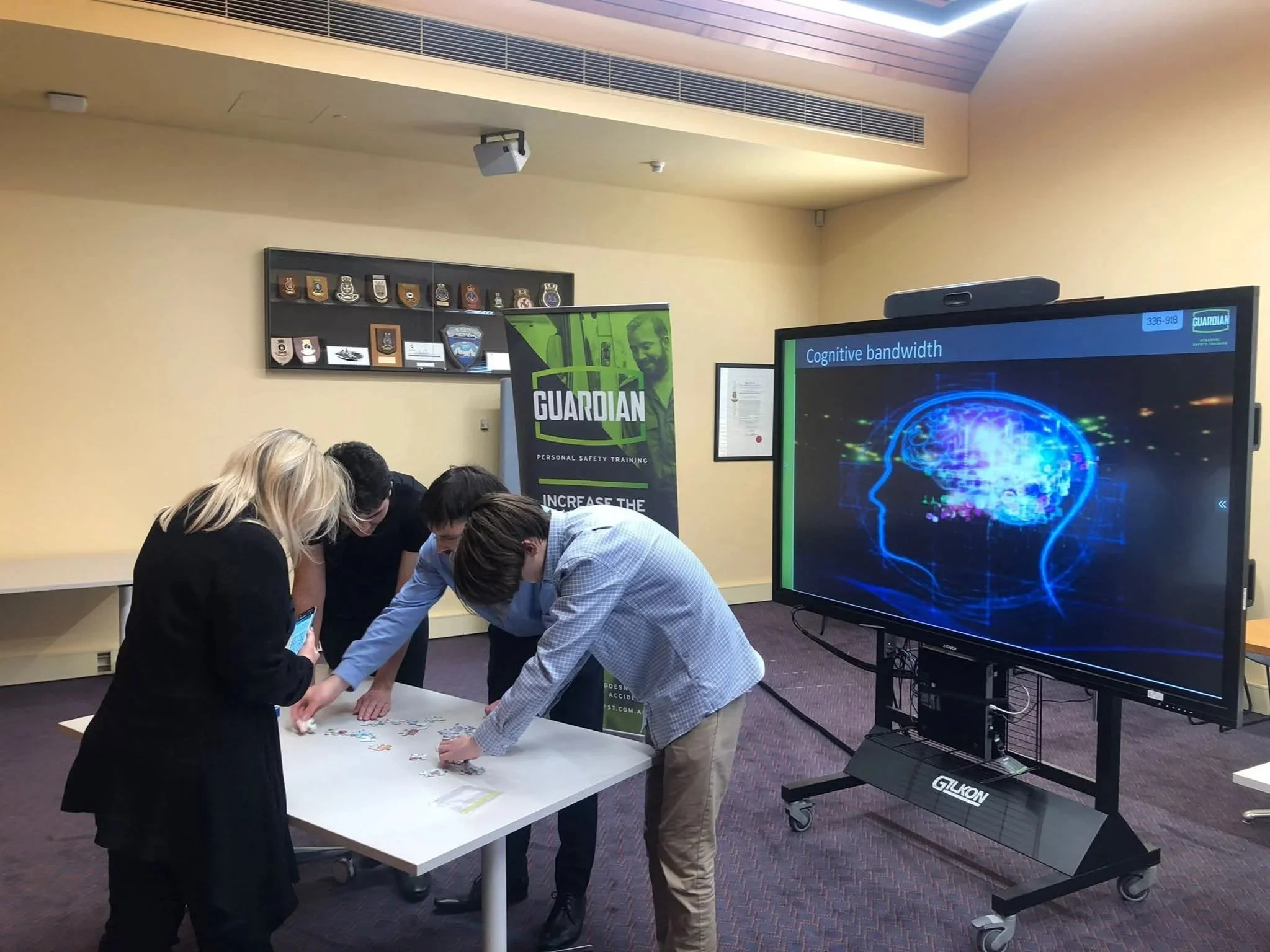 Group of people working together on a project at a table in a conference room. There is a large screen displaying a digital brain graphic and a banner with the Guardian logo and text 'Personal Safety Training'.