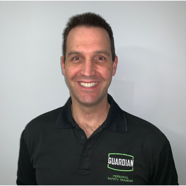 Smiling man wearing a black polo shirt with 'GUARDIAN Personal Safety Training' logo, standing against a plain wall.